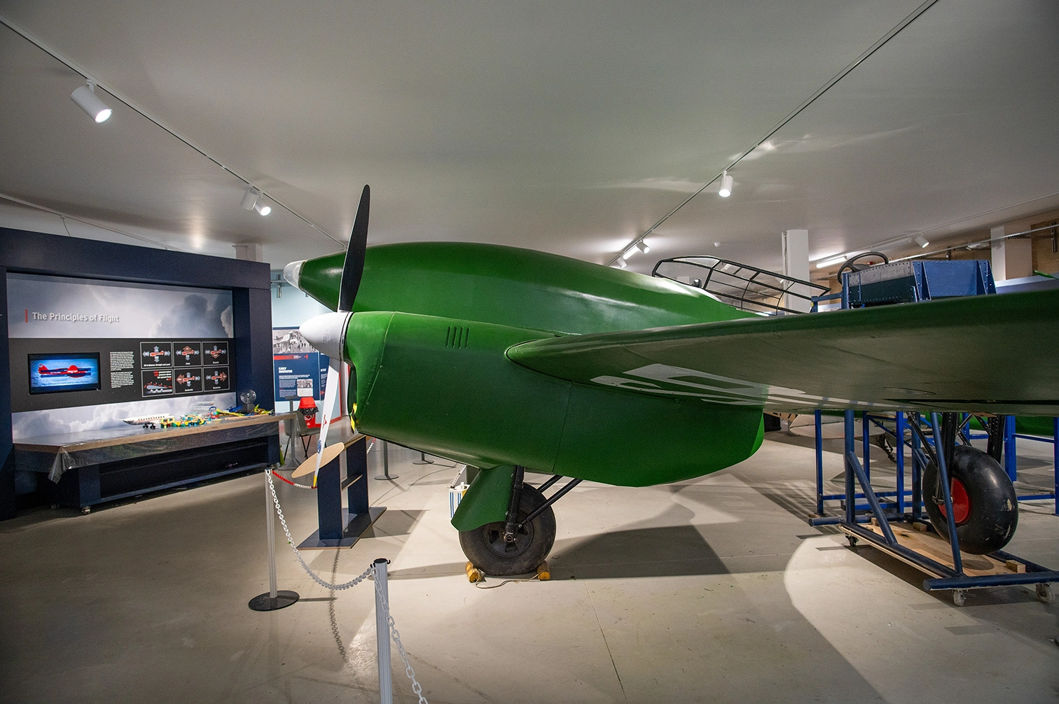 The image shows a green vintage aircraft displayed in an indoor museum setting. The aircraft is positioned on a stand with barriers around it. In the background, there is an exhibit titled 'The Principles of Flight' featuring informational displays and models. The lighting is bright, highlighting the details of the aircraft and the exhibit area.