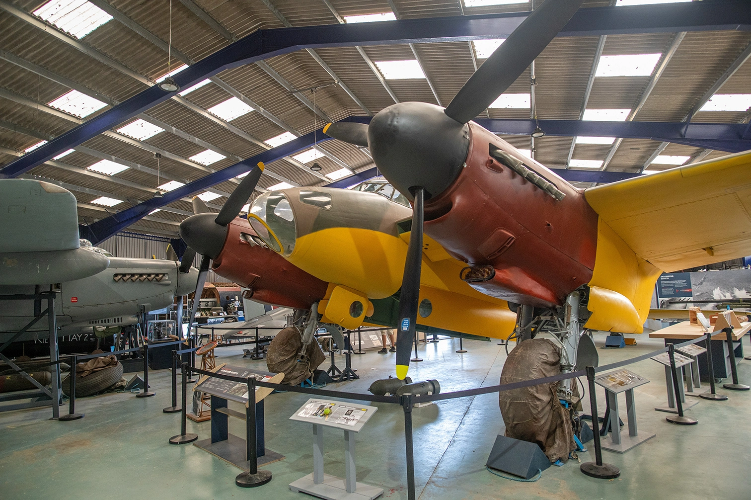 The image depicts an indoor aviation museum with several aircraft on display. The central focus is a brightly colored aircraft with a yellow nose and red body, featuring a prominent propeller. Other aircraft and exhibits are visible in the background, with informational stands and barriers set up around the displays. The setting is well-lit with a high ceiling and industrial lighting.
