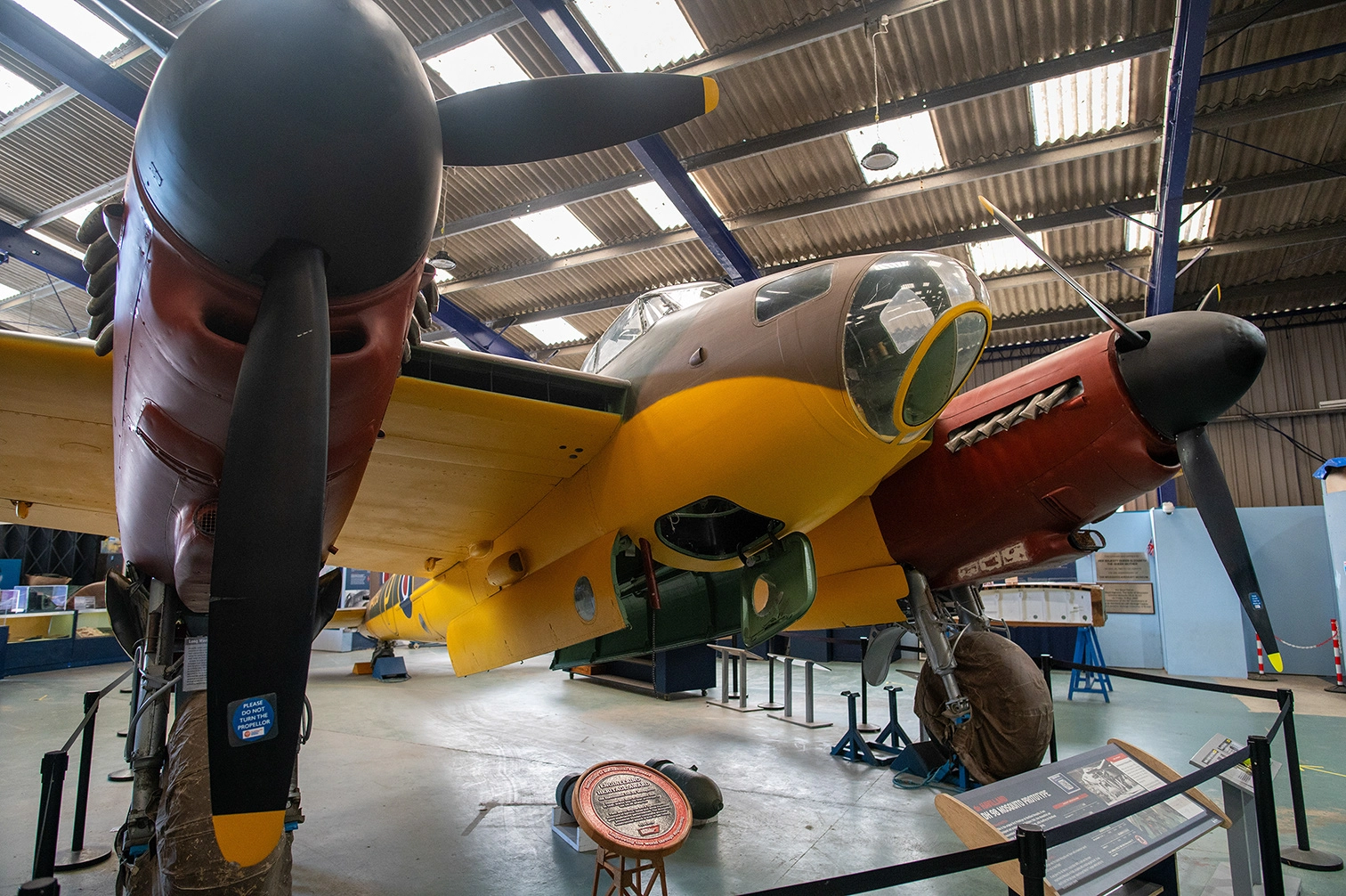 The image depicts a vintage military aircraft displayed in a museum setting. The aircraft is primarily yellow with a red nose and has a large propeller. It is housed in a spacious hangar with other exhibits visible in the background. Informational displays and barriers are set up around the aircraft for visitors.