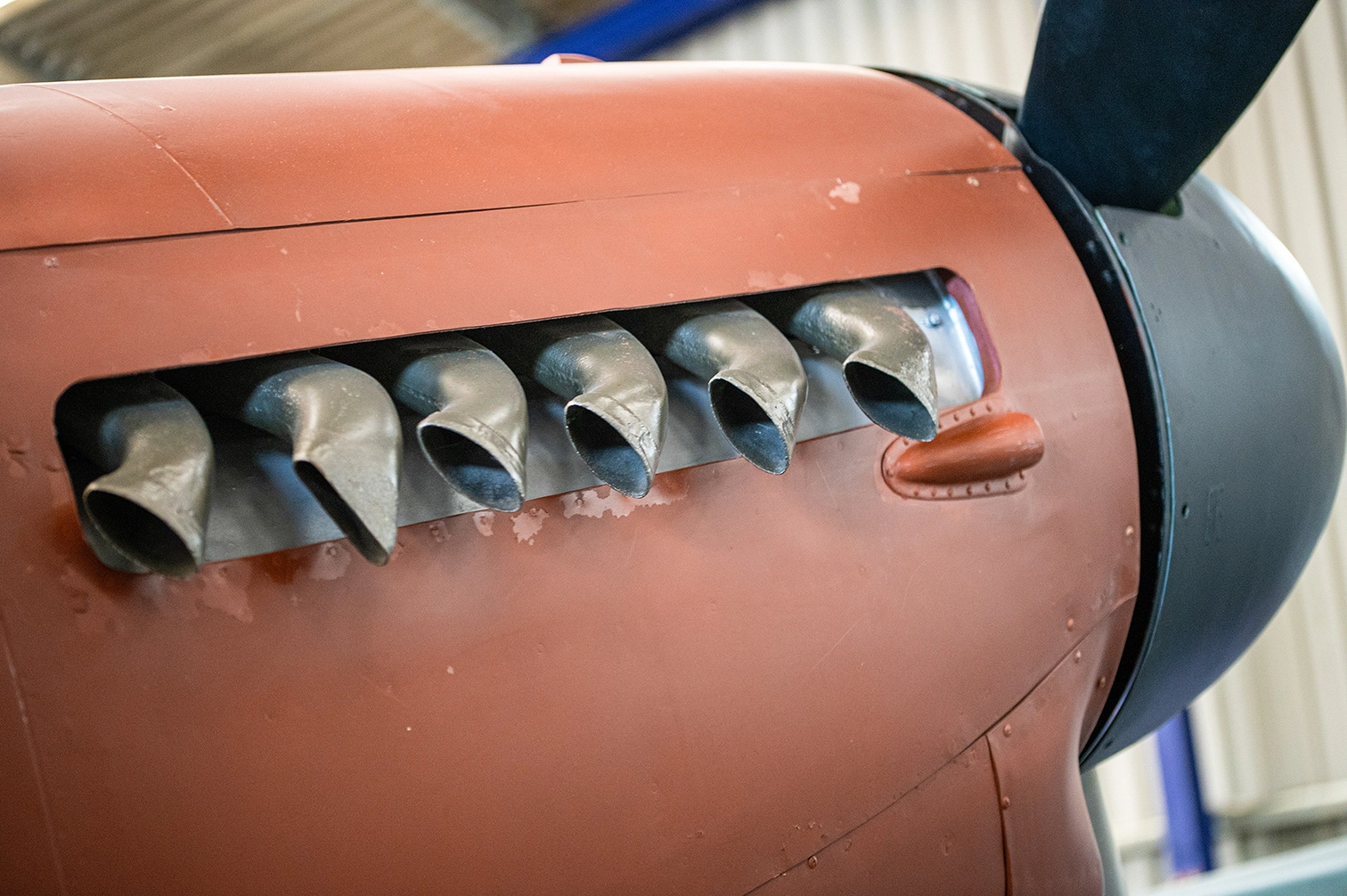 The image shows a close-up view of an aircraft engine with a propeller. The engine has a reddish-brown body with six exhaust pipes visible. The propeller has a dark, possibly black or dark blue, finish. The aircraft appears to be stationary, possibly in a museum or hangar setting.