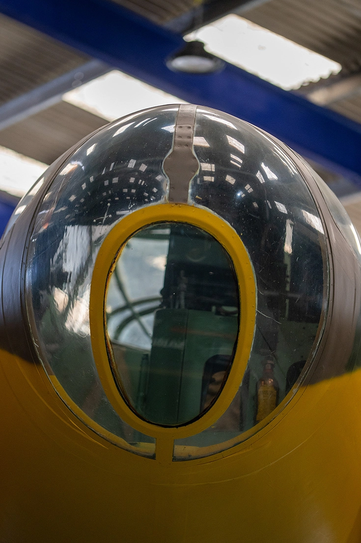 The image depicts the front section of an aircraft, specifically focusing on the transparent nose cone. The nose cone is framed with a yellow ring and provides a view into the interior of the aircraft, revealing parts of the cockpit and instrumentation. The reflective surface of the cone shows some distorted reflections of the surroundings, including overhead lighting and structural elements.