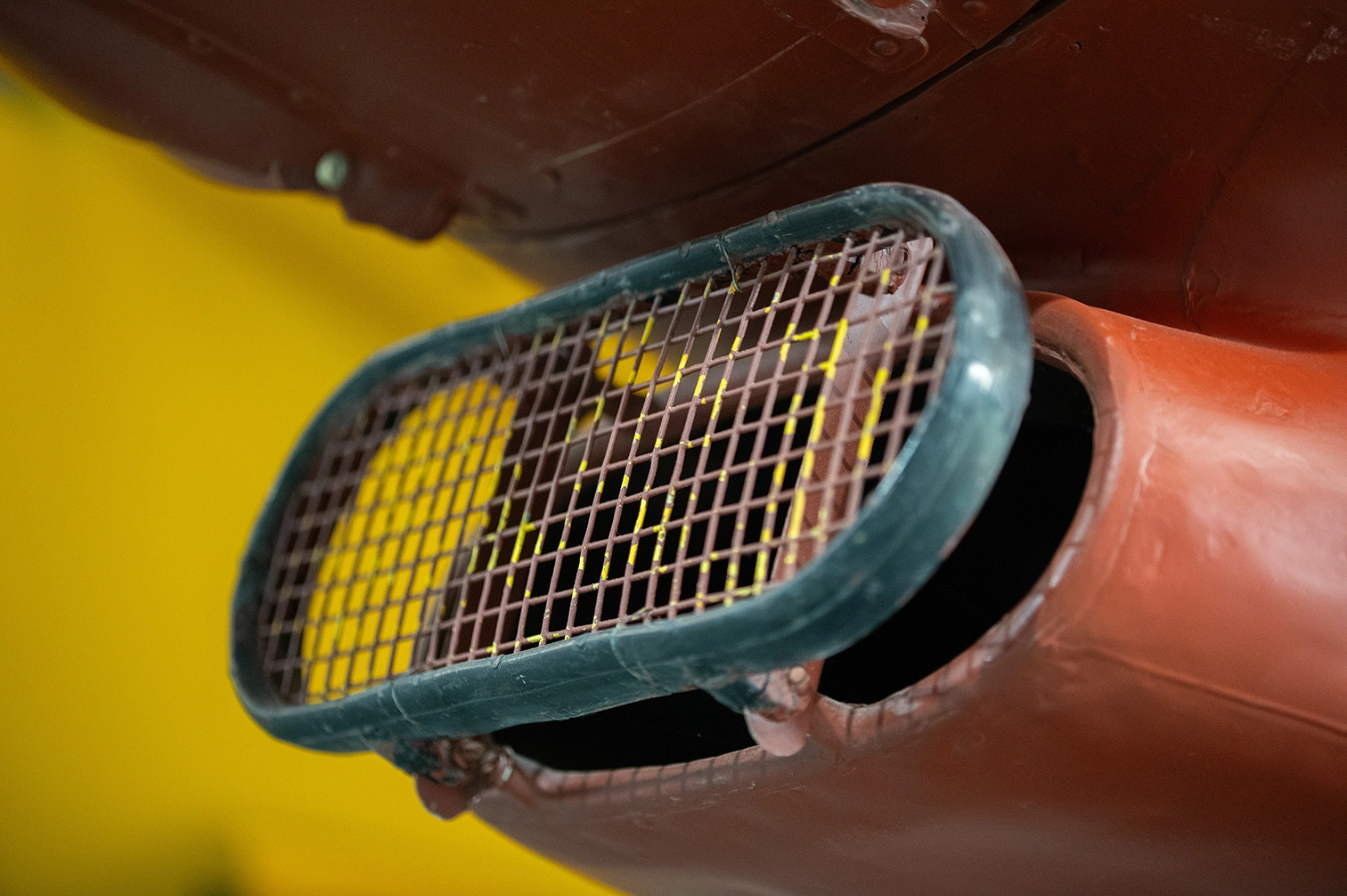 The image shows a close-up view of a mesh-covered vent or exhaust pipe, likely part of an engine or machinery. The mesh is slightly discolored, possibly due to heat or age, and the surrounding metal is painted in a combination of yellow and reddish-brown colors. The vent appears to be designed to allow airflow while preventing debris from entering.