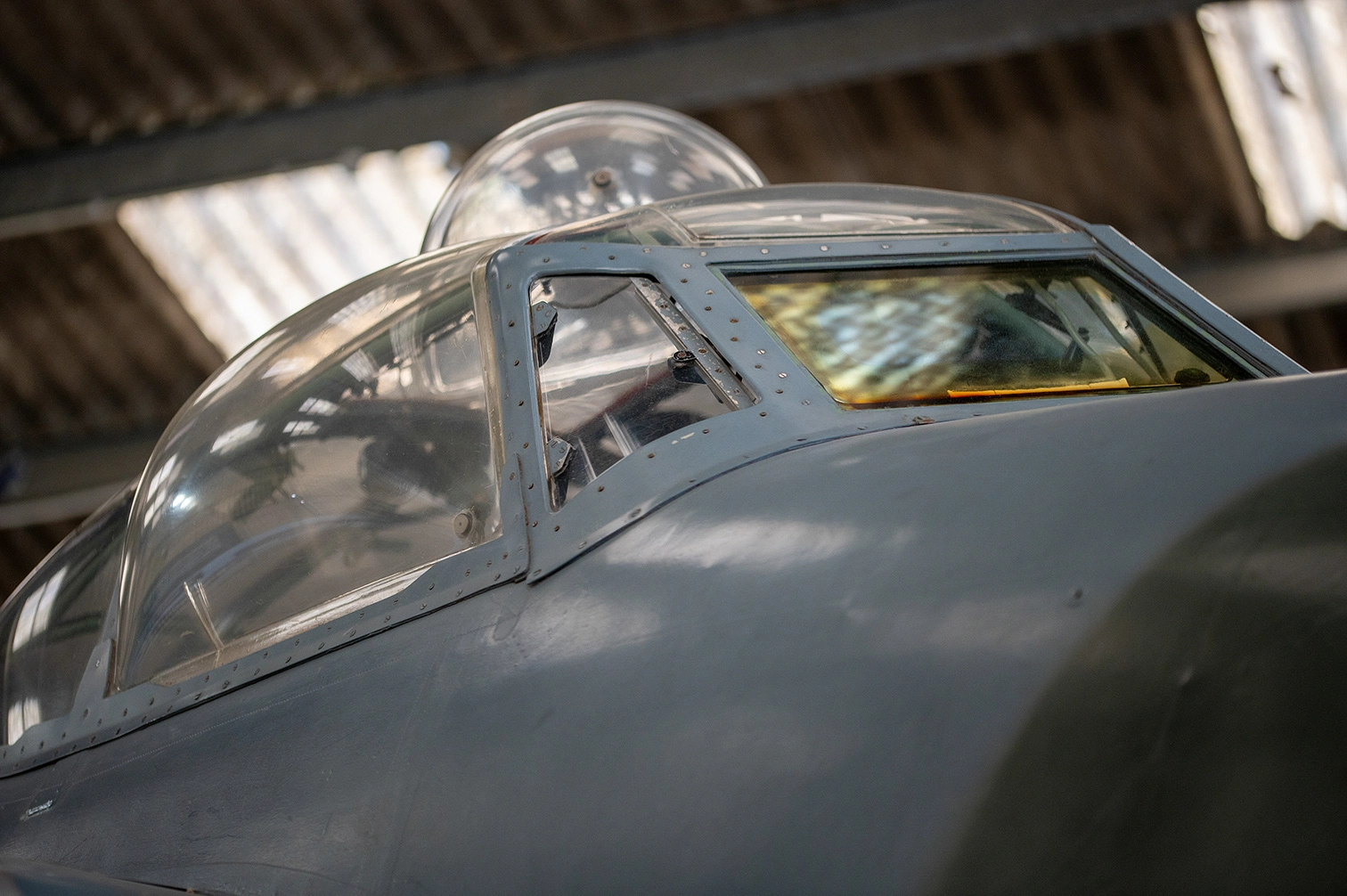 The image shows the cockpit of an aircraft with a clear canopy and metal frame. The aircraft is likely housed in a hangar or storage facility, as indicated by the background structure. The cockpit appears to be of an older model, possibly a vintage or historical aircraft. The lighting suggests it is daytime, with natural light illuminating the scene.