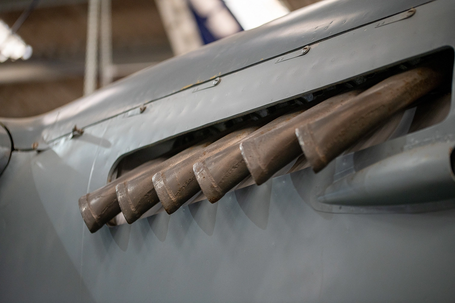 The image shows a close-up view of an aircraft, focusing on the exhaust pipes of the engine. The aircraft appears to be an older model, possibly from the early to mid-20th century, given the design and materials used. The exhaust pipes are prominently displayed, indicating a powerful engine setup. The surrounding structure and materials suggest a robust and durable construction typical of vintage aircraft.