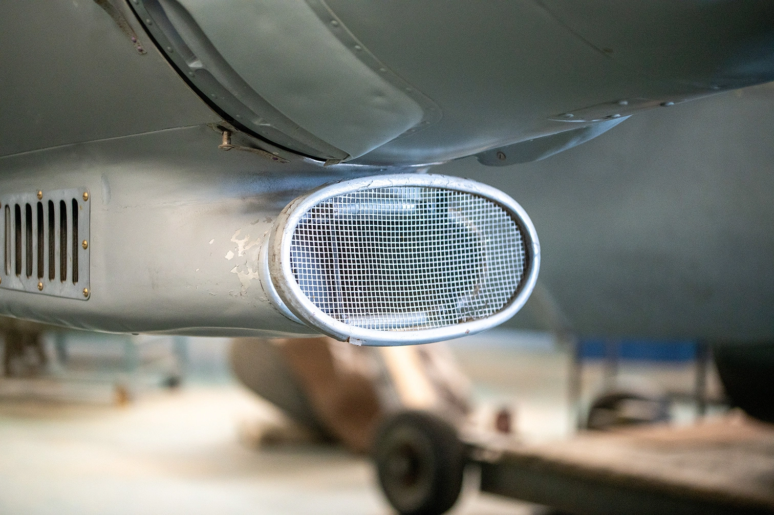 The image shows a close-up view of the underside of an aircraft, focusing on the exhaust pipe and a mesh-covered opening. The aircraft's metal surface shows signs of wear and some paint chipping. The background is blurred, indicating the aircraft is likely in a hangar or maintenance area. The overall scene suggests an environment of aircraft maintenance or inspection.