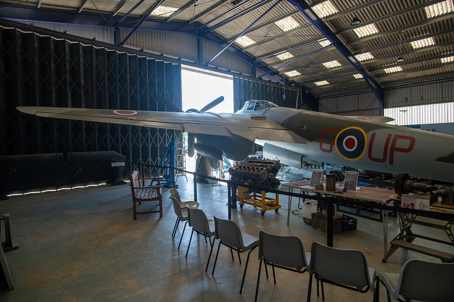 The image depicts an indoor setting, likely a museum or hangar, featuring a vintage military aircraft. The aircraft is prominently displayed with its wingspan extending across the space. The fuselage bears the markings 'GOUP' and a circular insignia. Several chairs are arranged in front of the aircraft, suggesting an area for presentations or briefings. The setting includes various informational displays and equipment related to the aircraft.