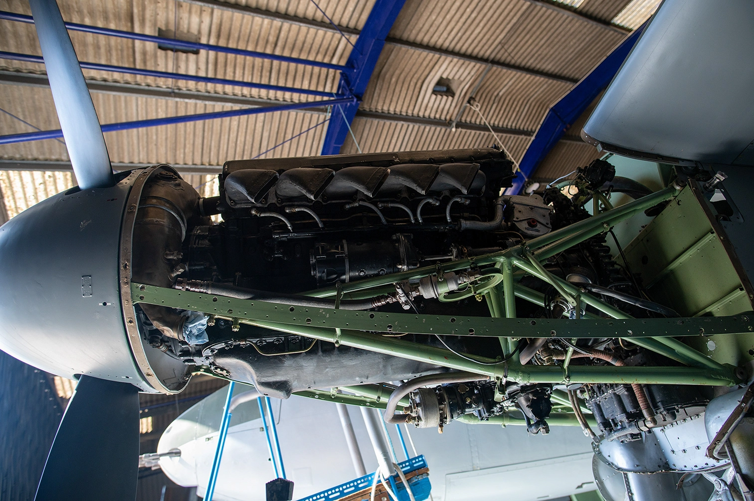 The image shows the front part of an aircraft, specifically focusing on the propeller and engine area. The aircraft is housed indoors, likely in a hangar, with natural light filtering through the windows in the background. The propeller has a polished, metallic finish, and the engine cowling appears to be made of metal with some signs of wear and rivets visible. The setting suggests that the aircraft might be on display or undergoing maintenance.
