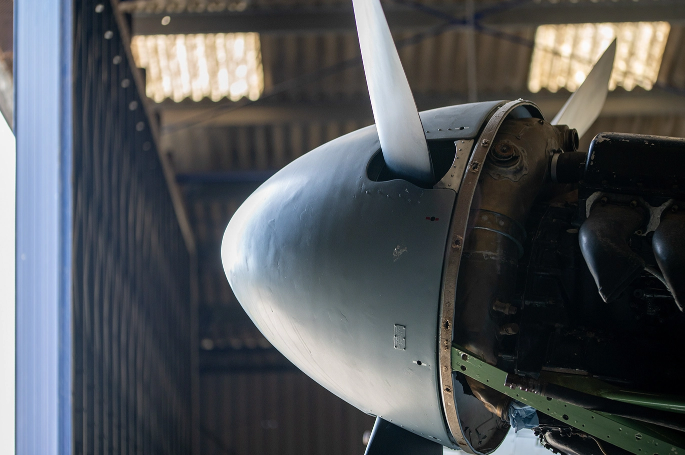 The image shows the front part of an aircraft, specifically focusing on the propeller and engine area. The aircraft is housed indoors, likely in a hangar, with natural light filtering through the windows in the background. The propeller has a polished, metallic finish, and the engine cowling appears to be made of metal with some signs of wear and rivets visible. The setting suggests that the aircraft might be on display or undergoing maintenance.