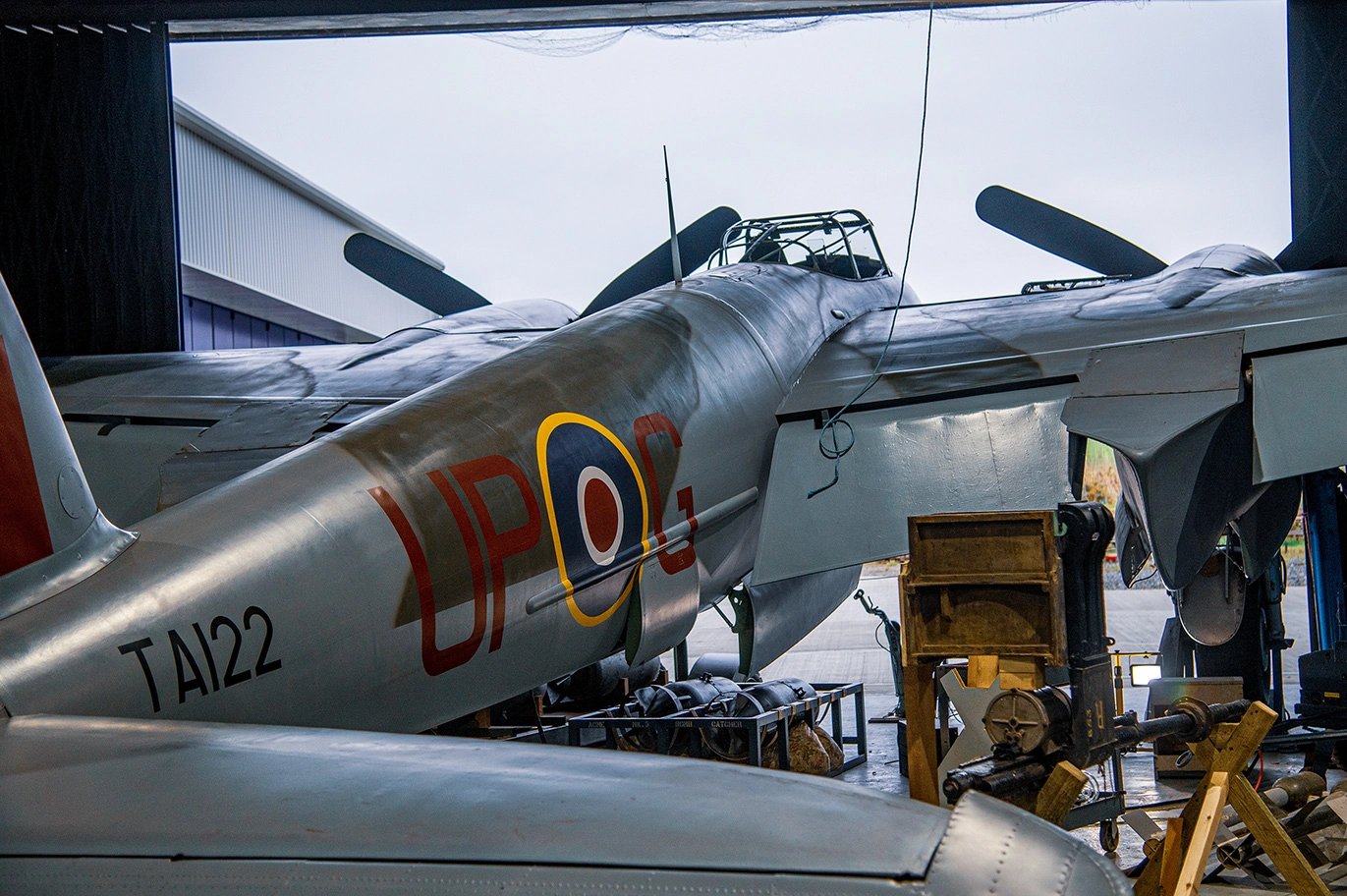 The image depicts a vintage military aircraft housed in a hangar. The aircraft has the markings 'UPO' and a roundel symbol on its fuselage, along with the tail number 'TA122'. The plane appears to be undergoing maintenance or restoration, as indicated by the surrounding equipment and scaffolding.