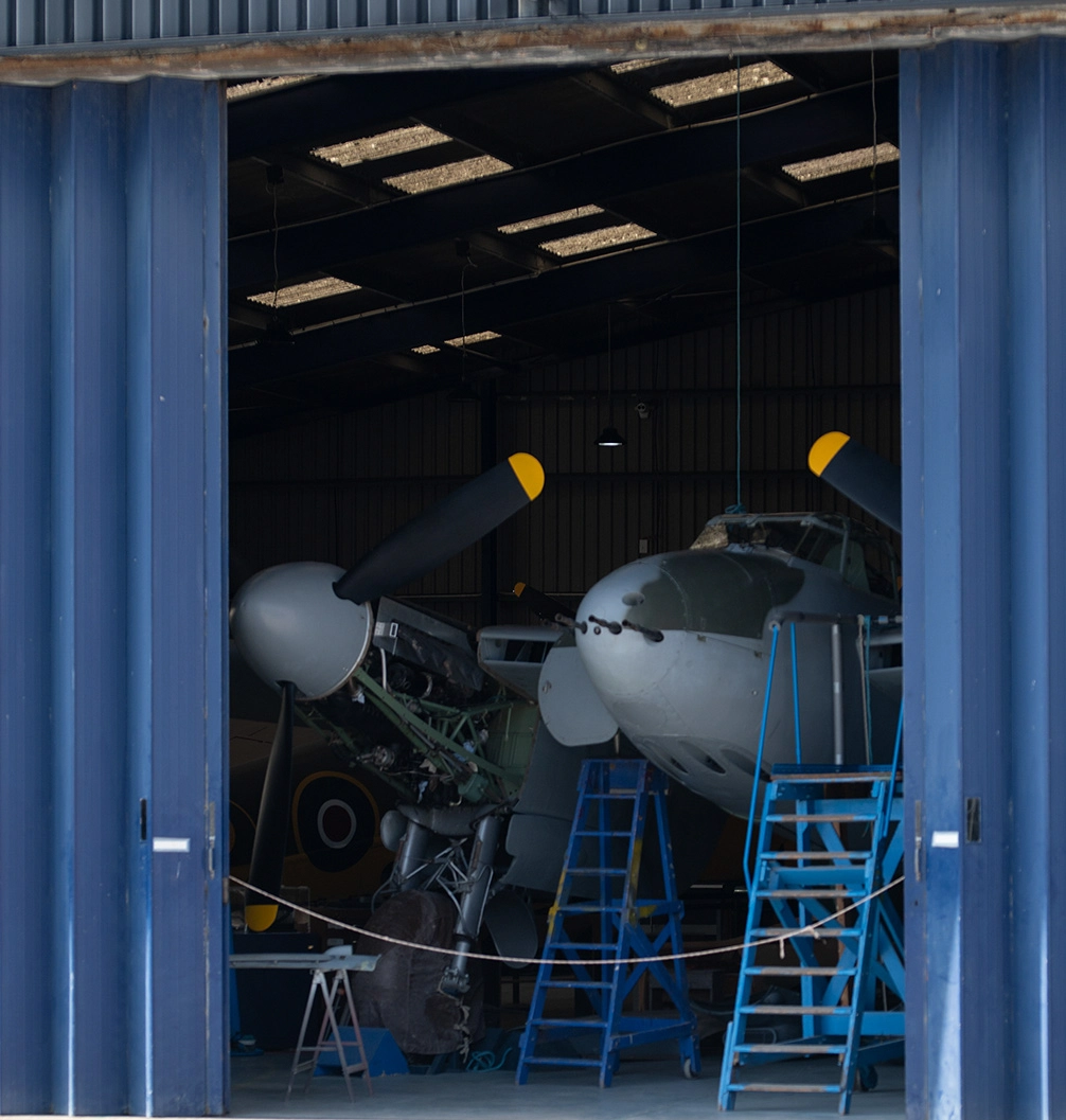 The image shows an aircraft inside a hangar. The aircraft is elevated on supports, with its nose pointed towards the viewer. The hangar has a corrugated metal exterior with blue doors, and the interior is dimly lit with overhead lights. There are ladders and other equipment around the aircraft, indicating maintenance or restoration work.