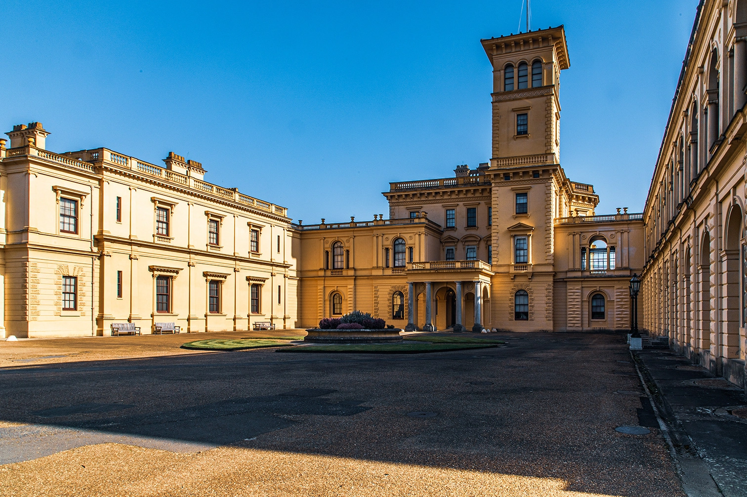 The image depicts a grand, historic building with classical architecture, featuring a prominent tower and a large courtyard. The structure is bathed in warm sunlight, highlighting its intricate details and symmetrical design. The building appears to be well-maintained, with a clean facade and neatly arranged windows. Benches and a small garden area are visible in the courtyard, adding to the serene and majestic atmosphere of the scene.
