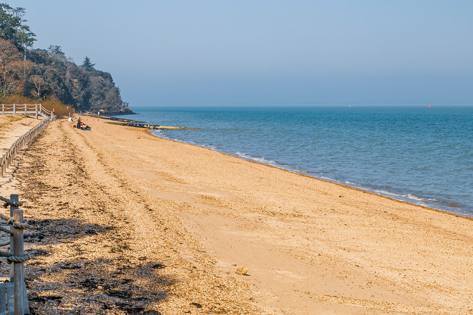 The image depicts a serene beach scene with a sandy shore extending into the distance. A wooden walkway leads from a paved path onto the beach, marked by a life preserver sign. In the background, there are trees, picnic tables, and small buildings, suggesting a recreational area. The sea is calm, and the sky is clear, indicating pleasant weather. The overall atmosphere is tranquil and inviting.