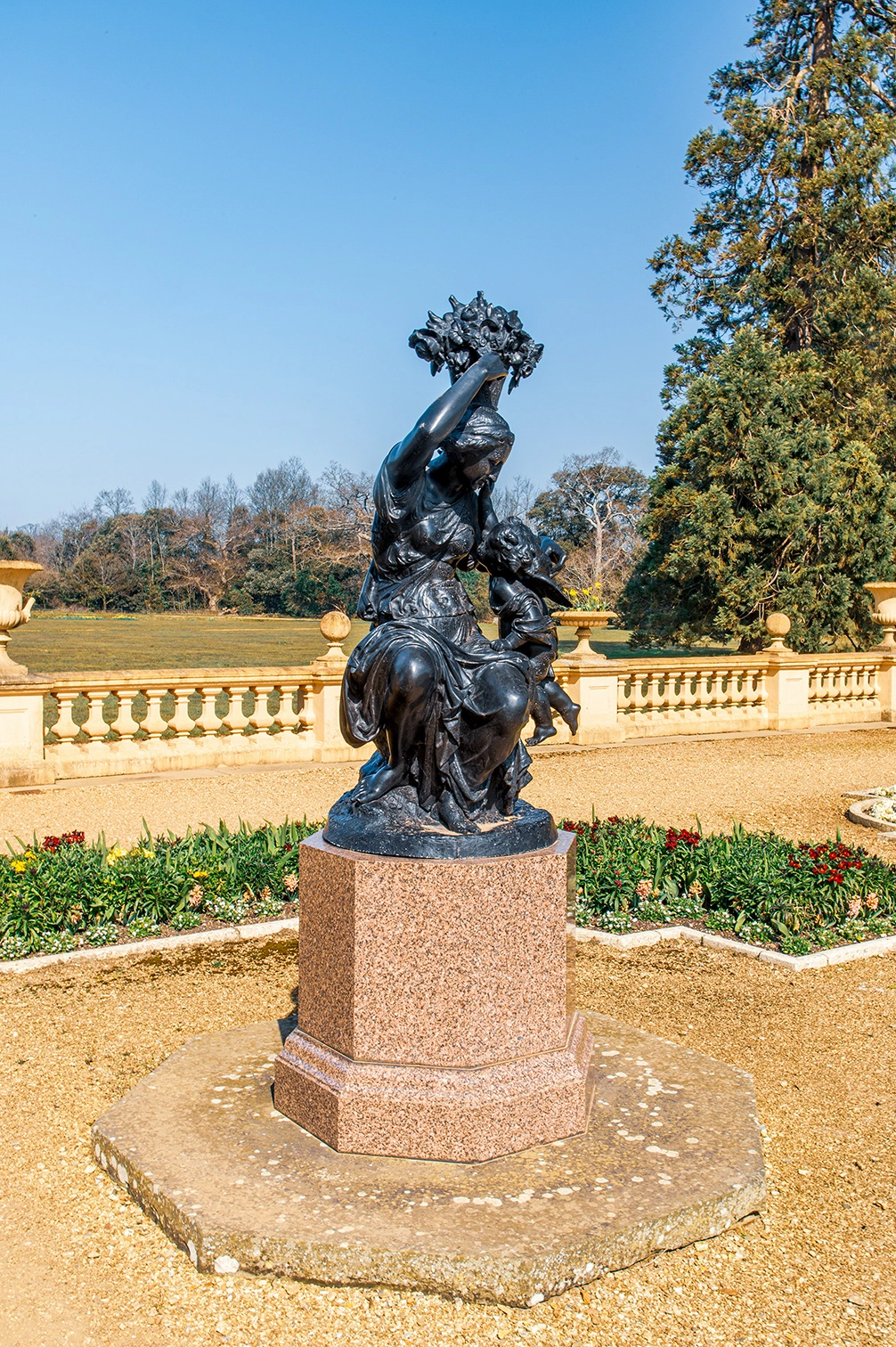 The image depicts a bronze statue situated in a well-maintained garden. The statue features two figures, one of which appears to be a woman holding a bunch of grapes above her head while the other figure, possibly a child, is reaching towards her. The statue is mounted on a granite pedestal with an octagonal base. The background includes a balustrade, lush greenery, and a clear blue sky.