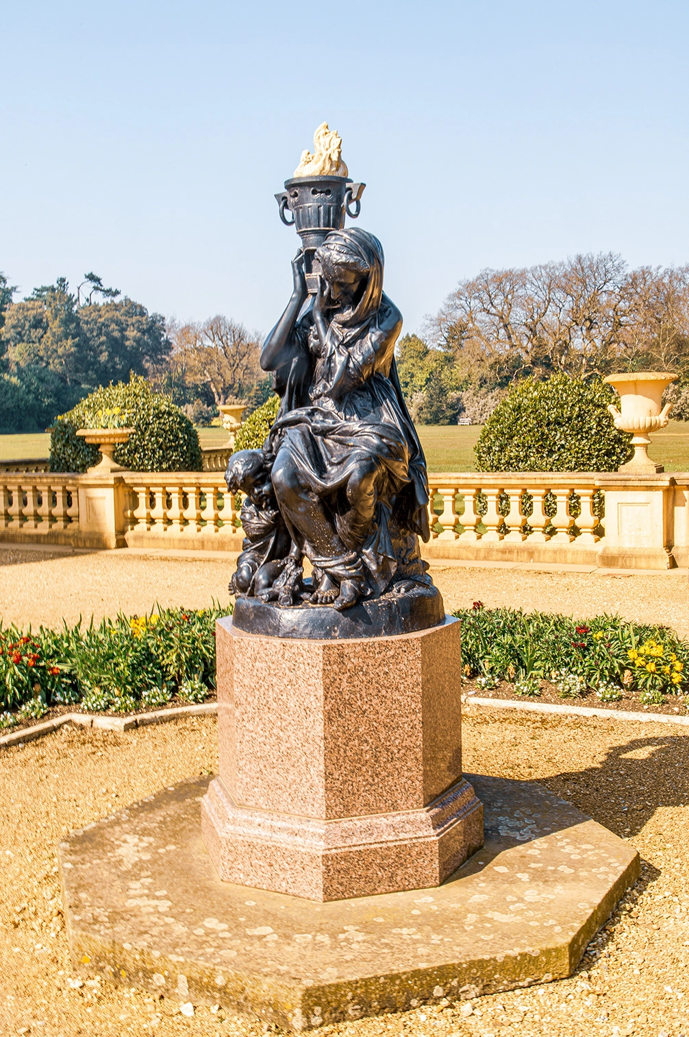 The image depicts a bronze statue situated in a landscaped garden. The statue features a robed figure holding a torch aloft, accompanied by a smaller figure. The statue stands on a granite pedestal, surrounded by a gravel path and manicured flower beds. In the background, there is a balustrade and more greenery, indicating a formal garden setting.