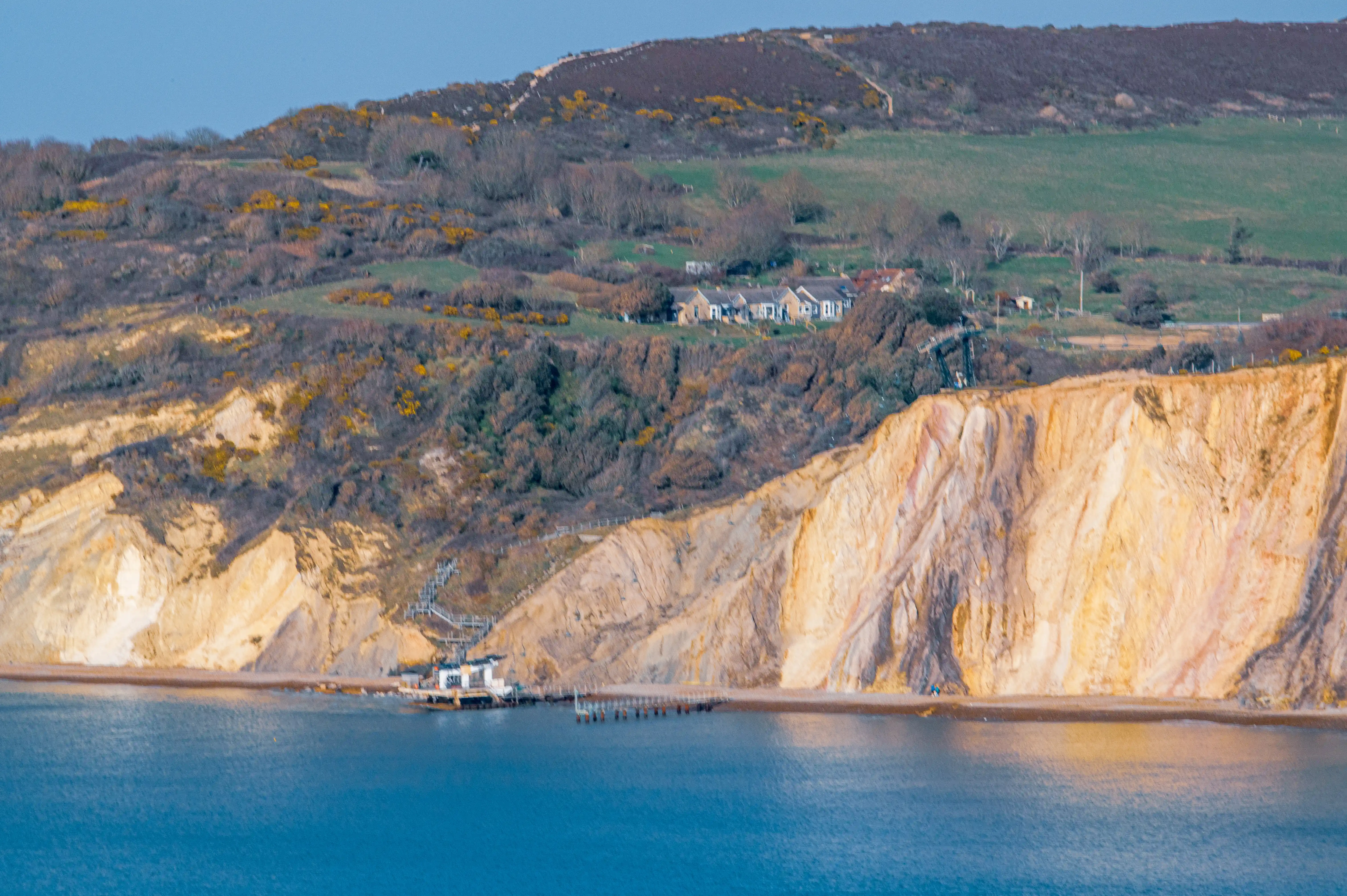 The image depicts a coastal landscape featuring prominent chalk cliffs, a beach, and a calm body of water. There are a few buildings situated on the hillside above the cliffs, surrounded by green fields and sparse vegetation. A pathway or staircase is visible descending the cliffs, leading to the beach area. The scene is tranquil, with gentle lighting suggesting either early morning or late afternoon.