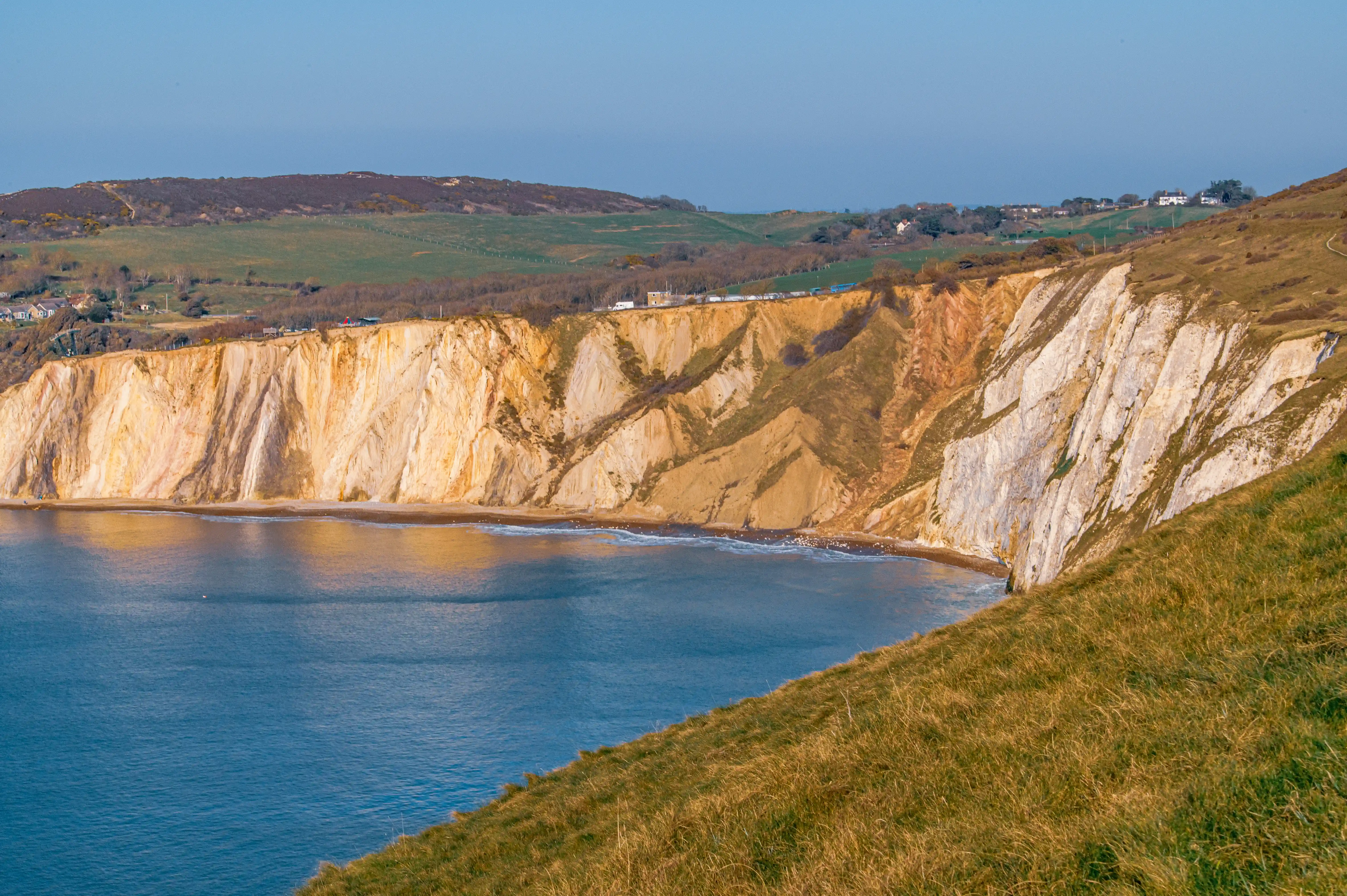 The image depicts a coastal landscape featuring steep, white cliffs that meet a calm body of water. The cliffs are layered with sedimentary rock, showcasing various shades of white and light brown. Above the cliffs, there is a rolling landscape of green hills and fields, dotted with a few buildings and houses. The scene is likely captured during the daytime, given the clear sky and the sunlight illuminating the cliffs and the water. The overall setting suggests a serene and picturesque natural environment.