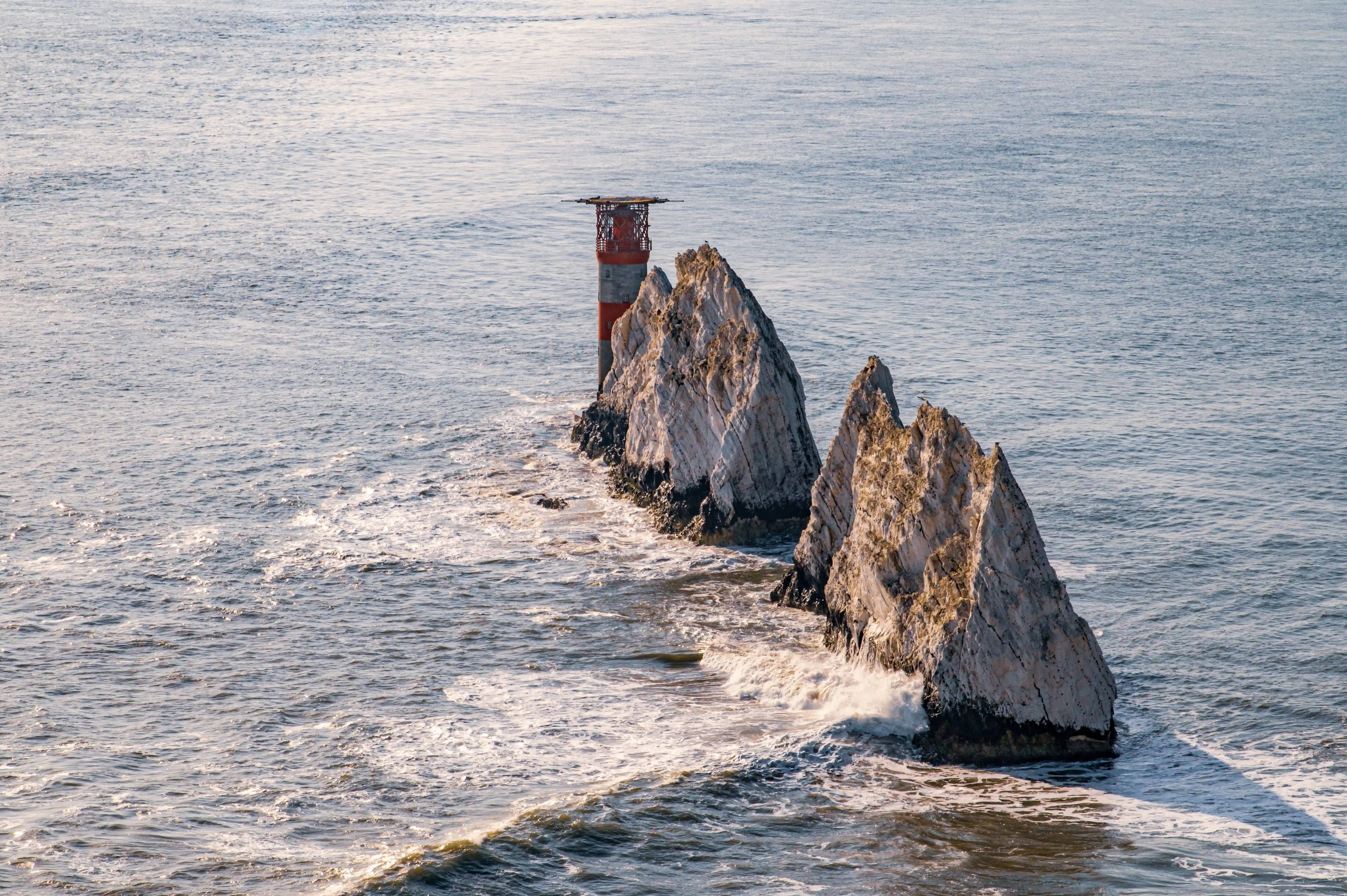 The image depicts a lighthouse situated on a rocky outcrop in the middle of the ocean. The lighthouse is red and white, standing prominently against the blue sea. The rocky formation consists of jagged, steep rocks that rise sharply from the water. Waves can be seen crashing against the base of the rocks, indicating a potentially rough sea. The overall scene is one of isolation and natural beauty, with the lighthouse serving as a crucial navigational aid in a challenging maritime environment.