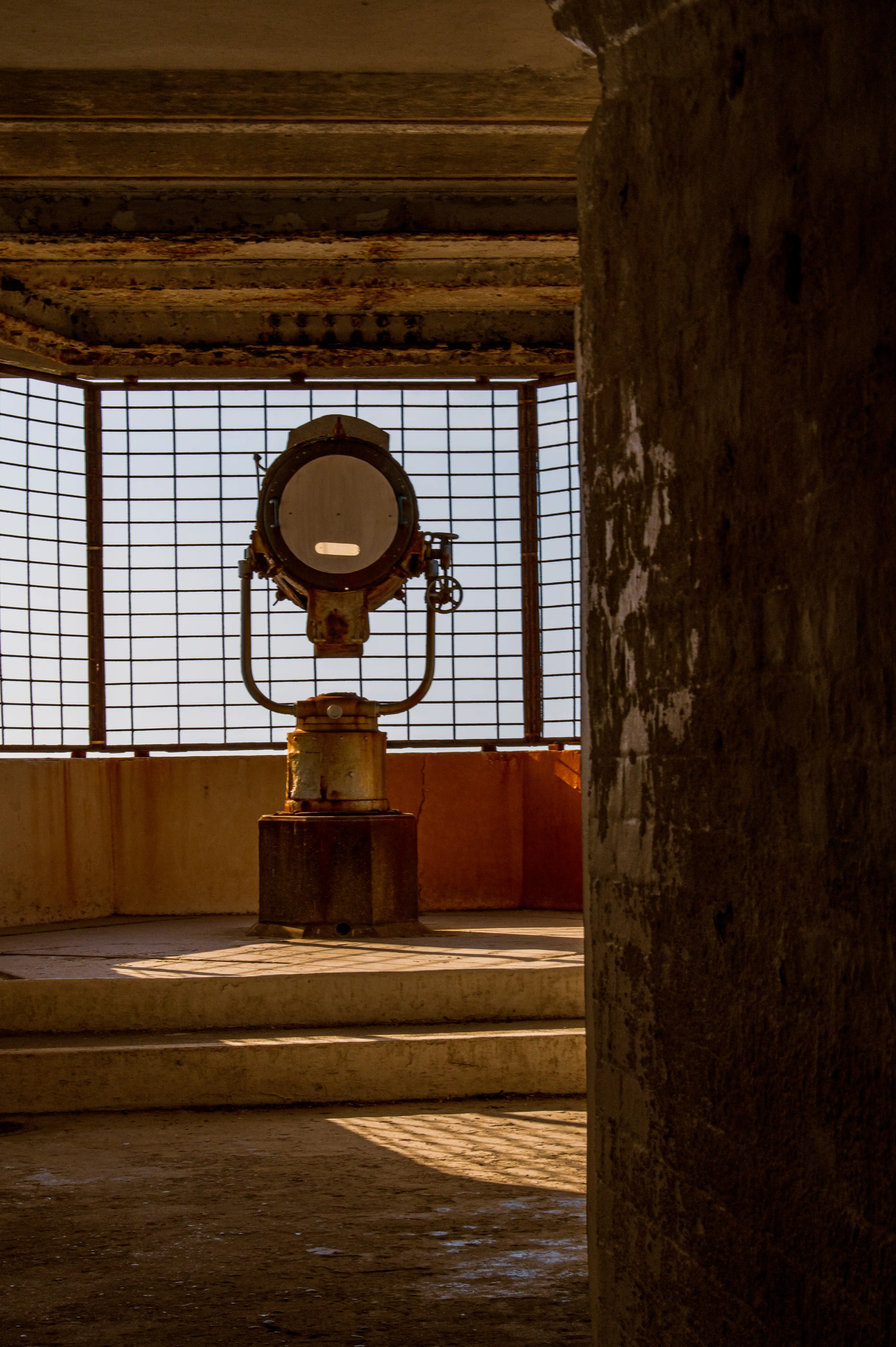 The image depicts an interior scene of what appears to be an old, possibly abandoned structure. A large searchlight or similar apparatus is centrally positioned, aimed towards a window with a grid pattern. The setting suggests an industrial or military context, with worn and weathered surfaces indicating age and disuse. Light streams in through the window, casting shadows and highlighting the textures of the walls and floor.