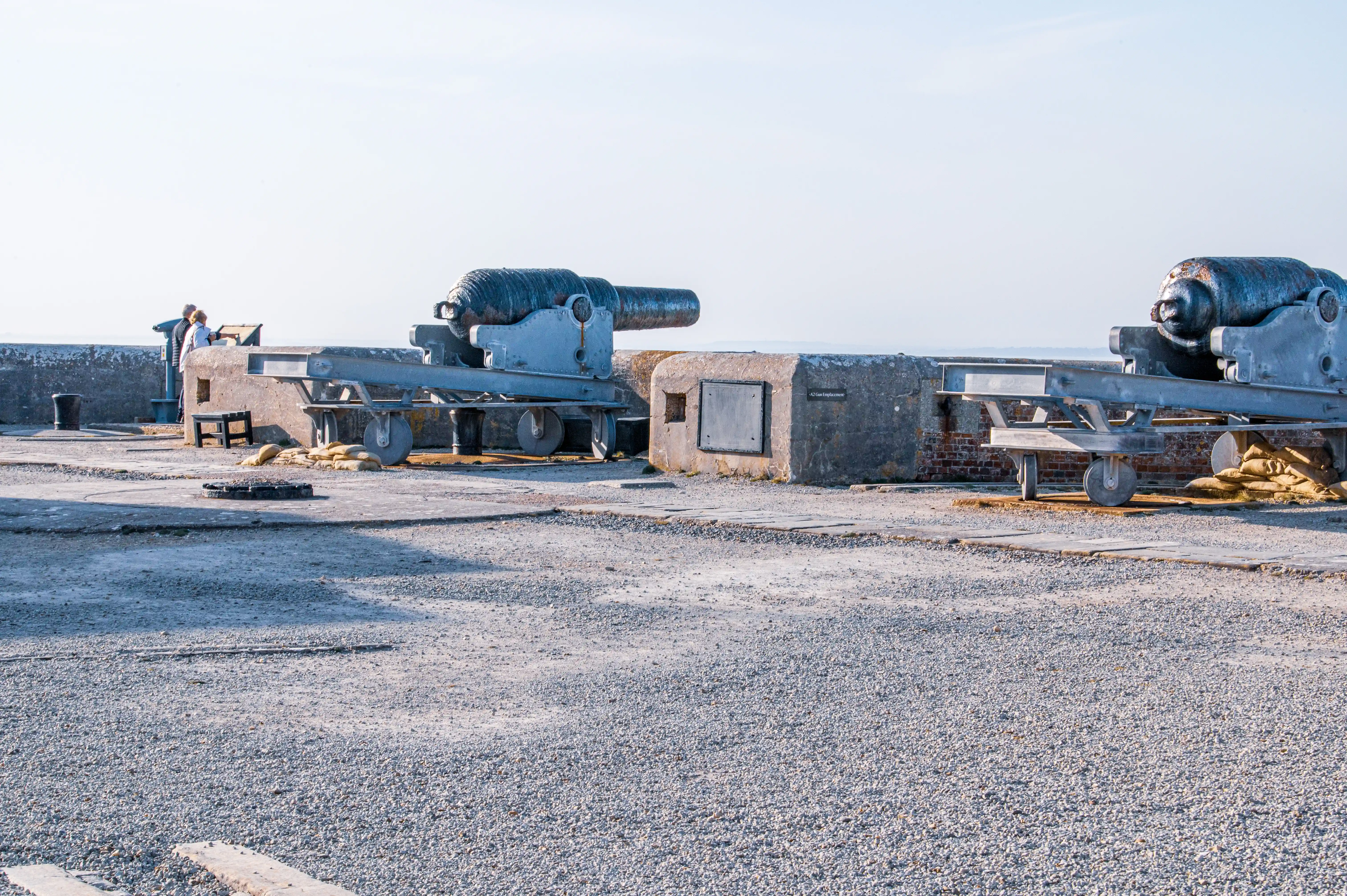 The image depicts a historical site featuring two large cannons mounted on metal carriages. The cannons are positioned on a gravel surface near a stone and brick structure, likely part of an old fort or battery. Two people are seen near the left cannon, possibly observing or taking photographs. The scene is set on a clear day with a bright sky, suggesting it is a well-preserved historical or tourist site.