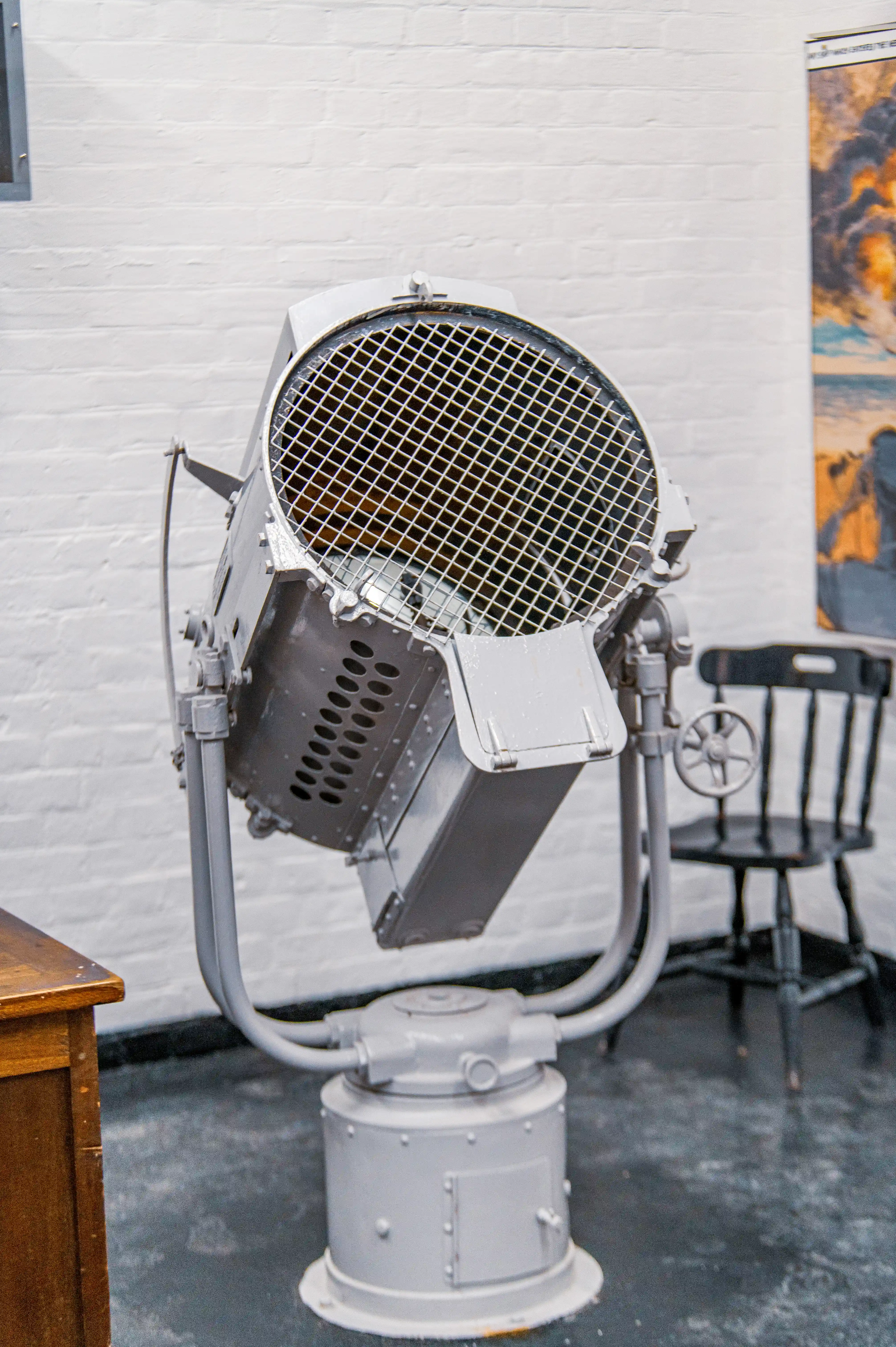 The image depicts a large, vintage searchlight mounted on a cylindrical base. The searchlight is positioned indoors, against a white brick wall. A wooden chair is visible to the right of the searchlight, and a wooden table is partially visible on the left. The setting appears to be a museum or historical display area.