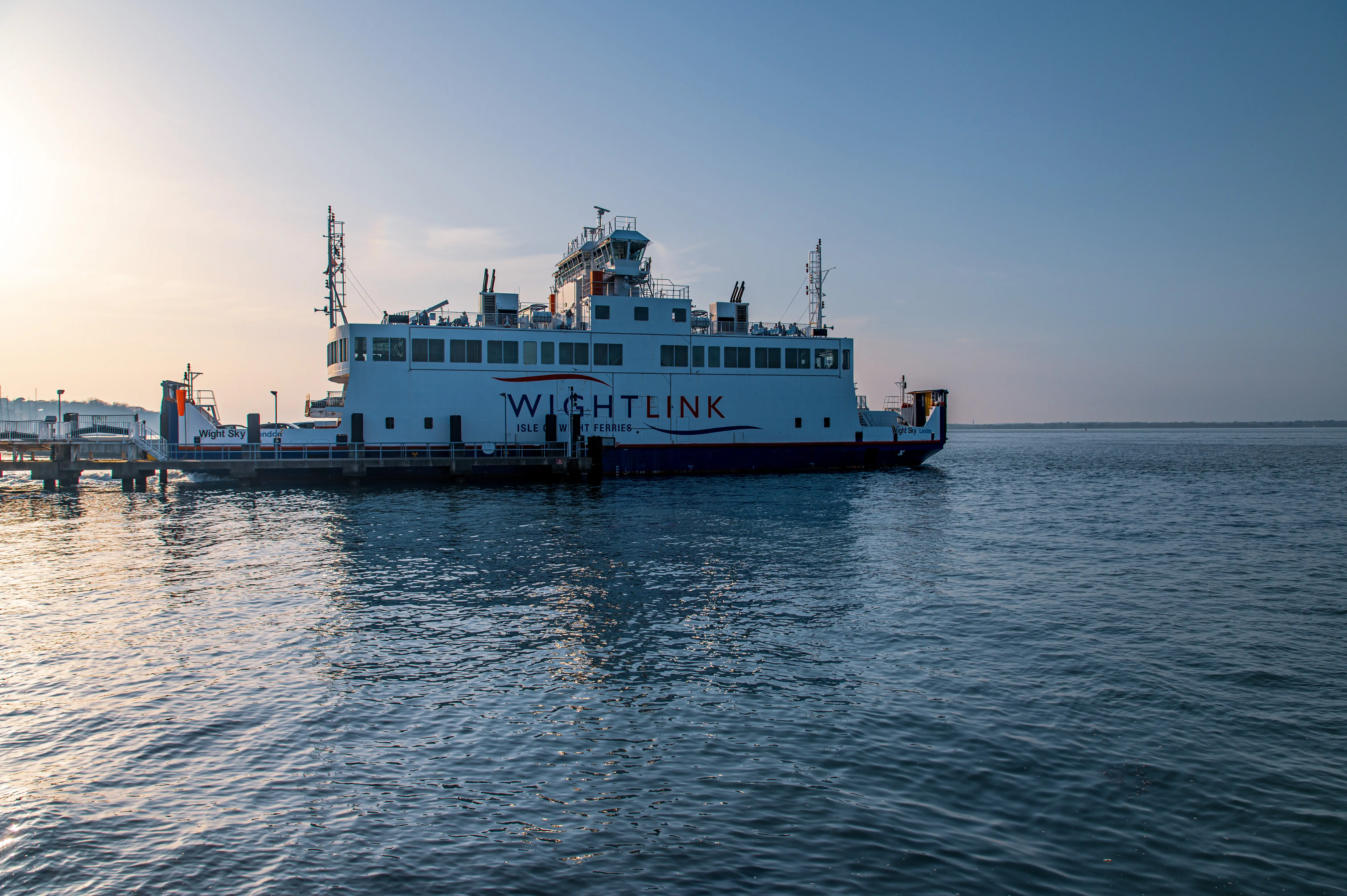 The image depicts a Wightlink ferry docked at a port during what appears to be either sunrise or sunset. The ferry is branded with the name 'Wightlink' and 'Isle of Wight Ferries.' The vessel is equipped with various antennas and equipment on its top deck, and it is connected to the dock via a boarding ramp. The water is calm, and the sky has a gradient of colors, indicating the time of day.