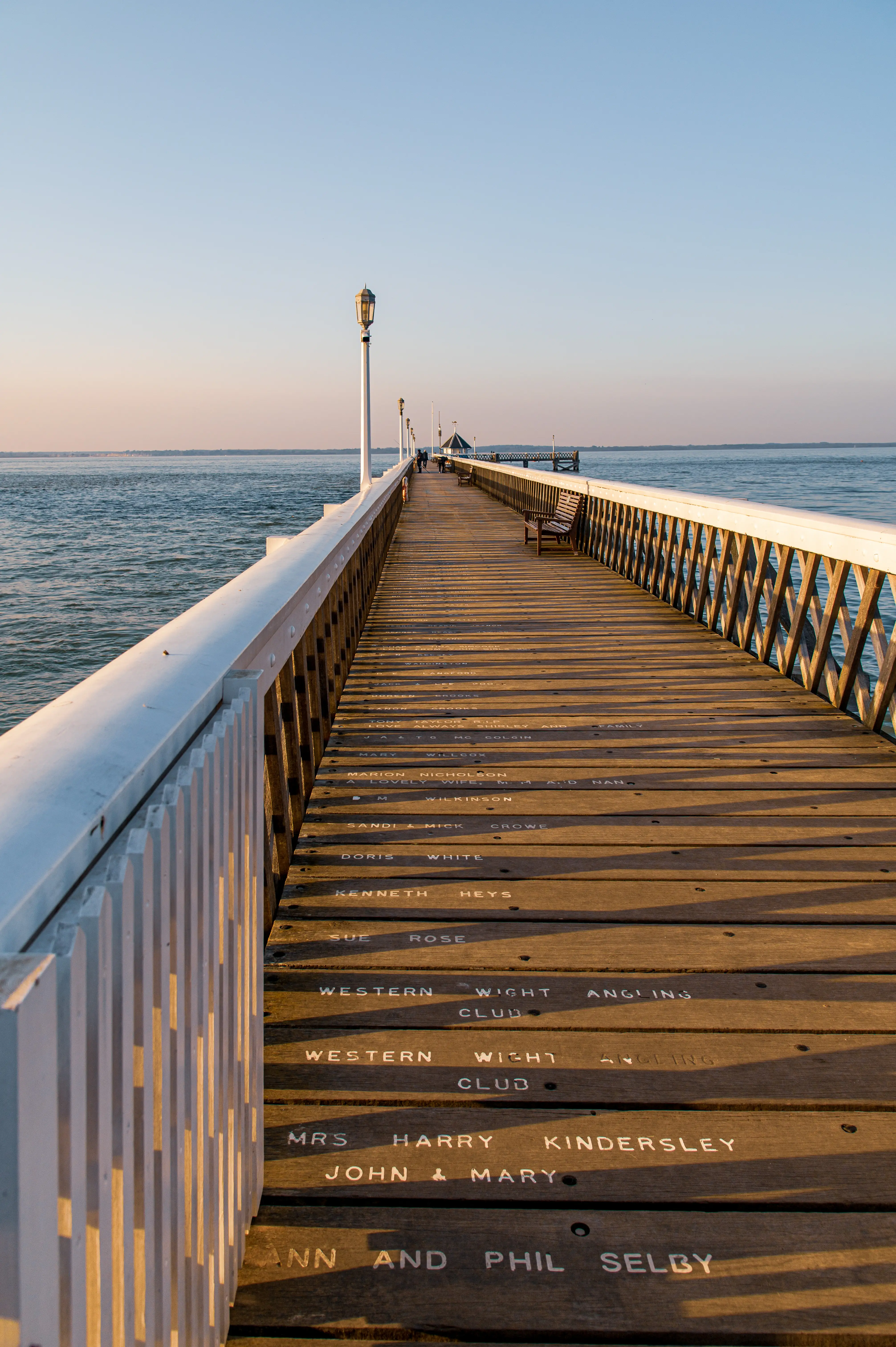 The image depicts a long wooden pier extending into a body of water, likely a sea or lake. The pier has a white railing on the left side and wooden benches along its length. Names and inscriptions are carved into the wooden planks of the pier. The scene is set during what appears to be either sunrise or sunset, with calm water and a clear sky in the background.