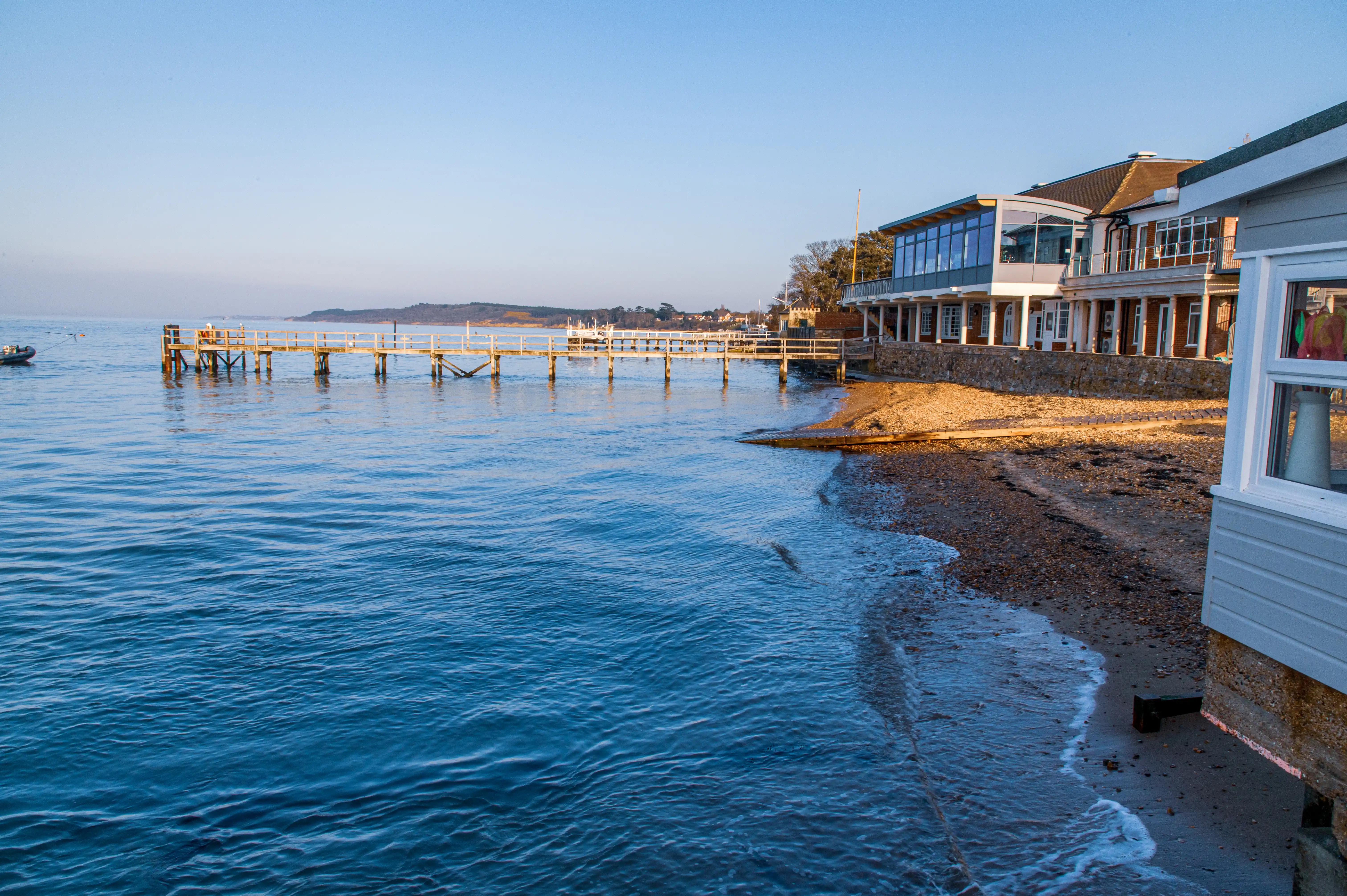 The image depicts a serene waterfront scene with a building complex situated along the shoreline. The complex includes modern architecture with large windows and a balcony, adjacent to a wooden pier extending into the calm water. The beach in the foreground has gentle waves lapping against the shore, and the overall atmosphere is tranquil with clear skies and distant landforms visible on the horizon.