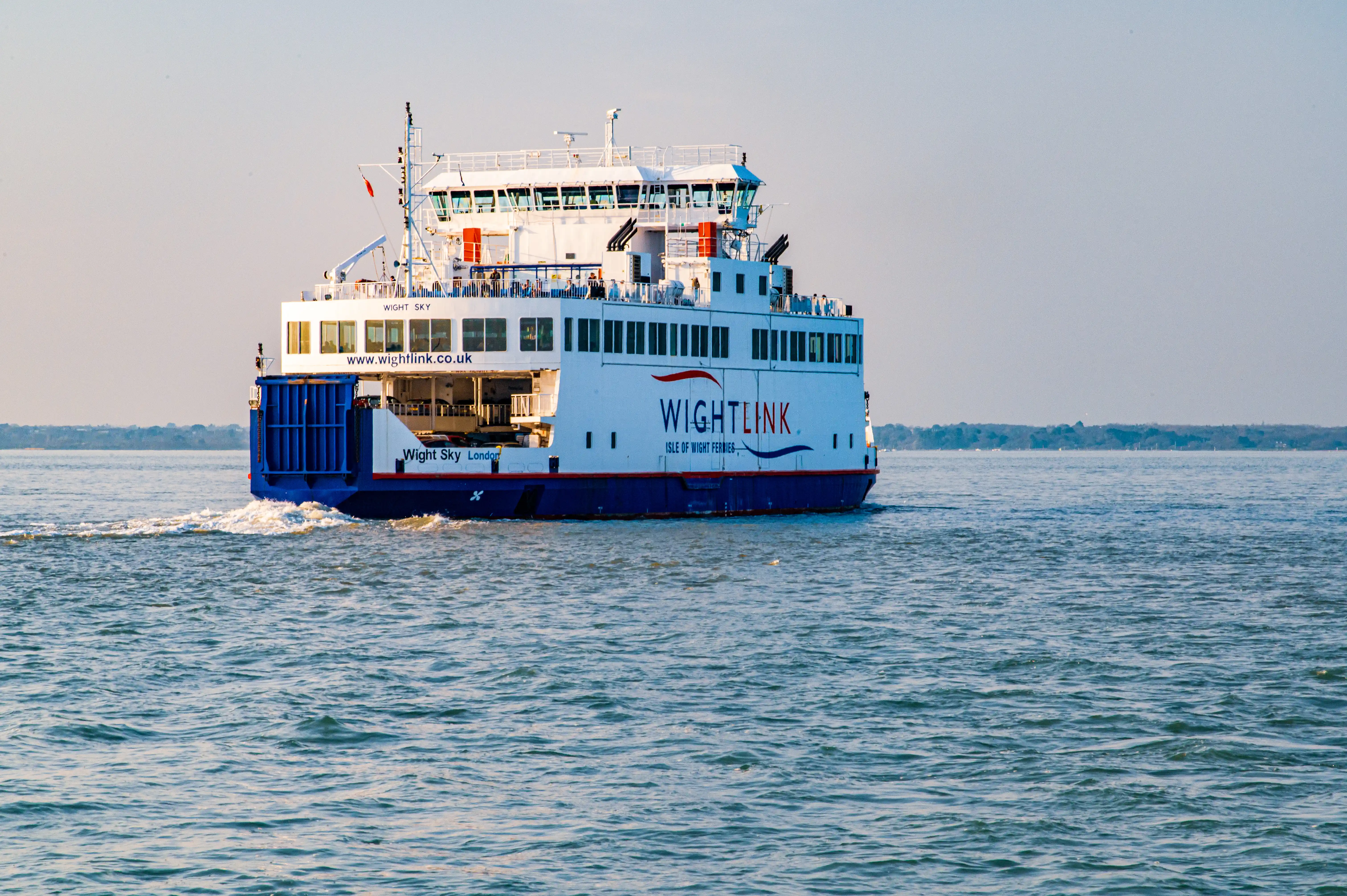 The image shows the Wightlink ferry, Wight Sky, sailing through calm blue waters. The ferry, predominantly white with a blue hull and red accents, displays WIGHTLINK ISLE OF WIGHT FERRIES on its side, along with the website www.wightlink.co.uk. The vessel is moving away from the viewer, creating a wake in its path. In the background, a tree-lined coastline is visible under a clear, pale sky.