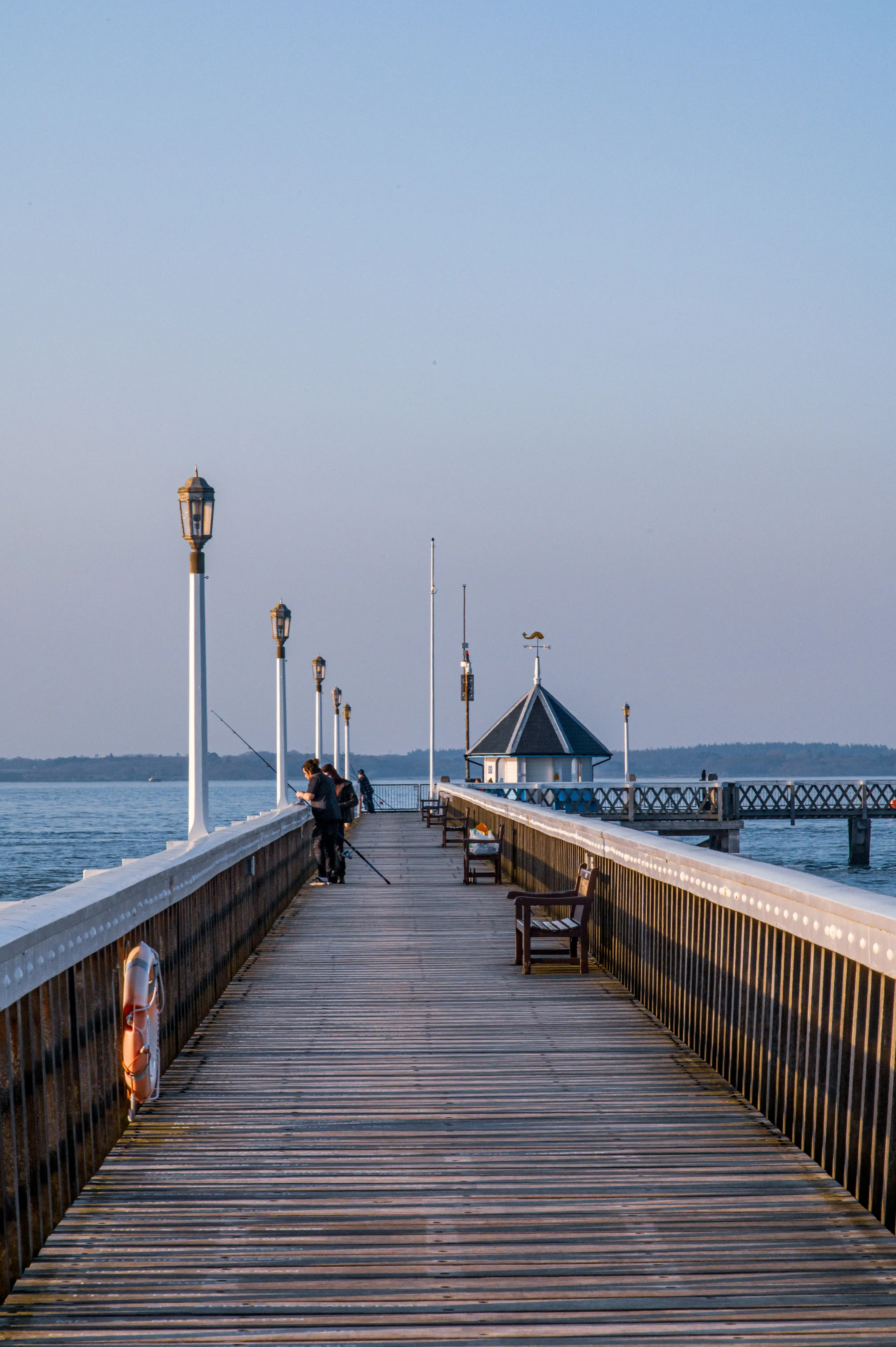 The image depicts a serene scene of a long wooden pier extending into a calm body of water. The pier is lined with vintage-style street lamps and has a few benches along its length. A small gazebo-like structure is visible at the end of the pier, and a few people are seen fishing. The sky is clear, and the overall atmosphere is tranquil and picturesque.