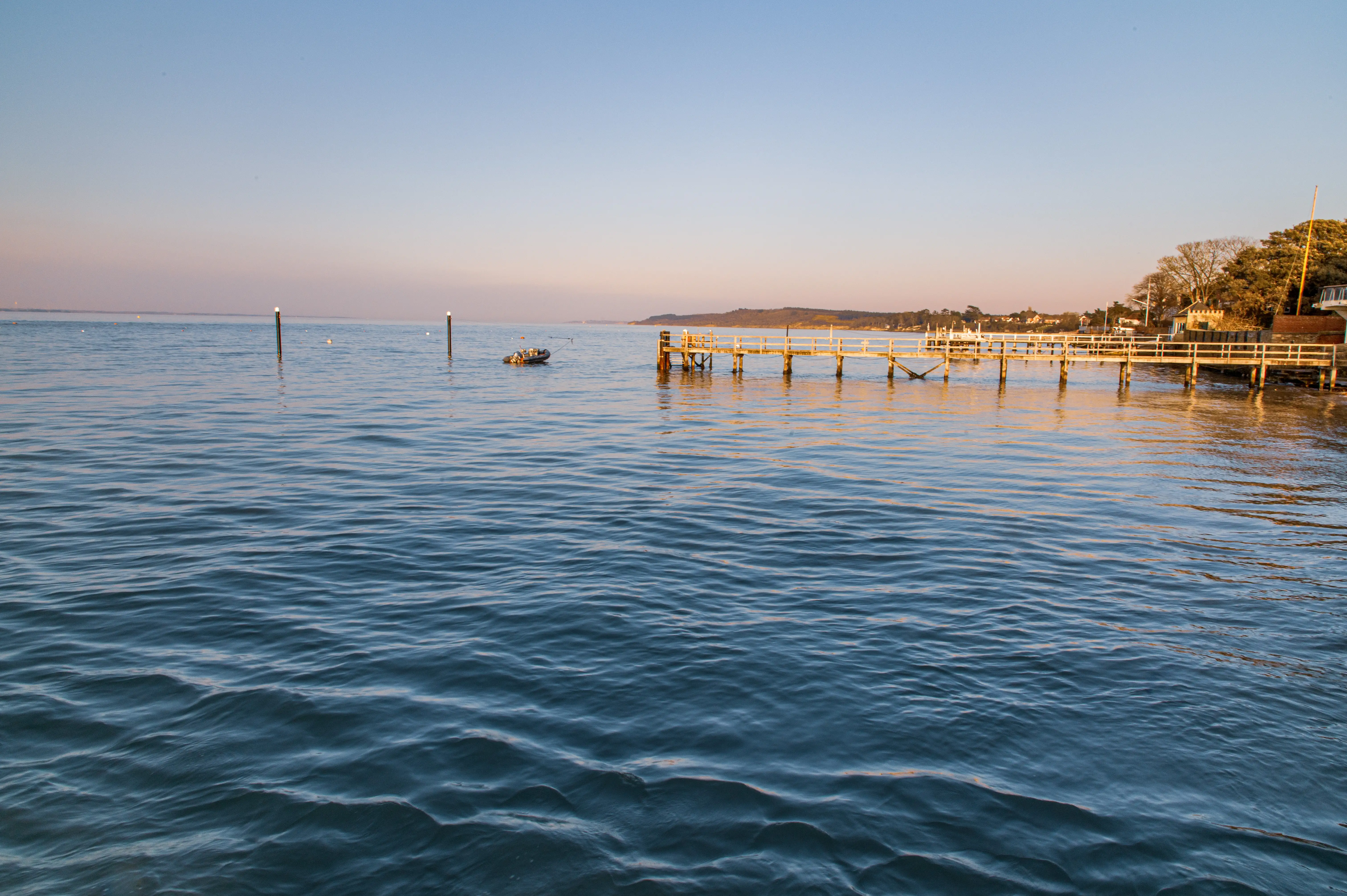 The image depicts a serene waterfront scene during what appears to be either sunrise or sunset. A wooden pier extends into calm waters, and a small boat is visible near the pier. The horizon shows a distant shoreline, and the sky is painted with soft hues of orange and blue. The overall atmosphere is tranquil and picturesque.