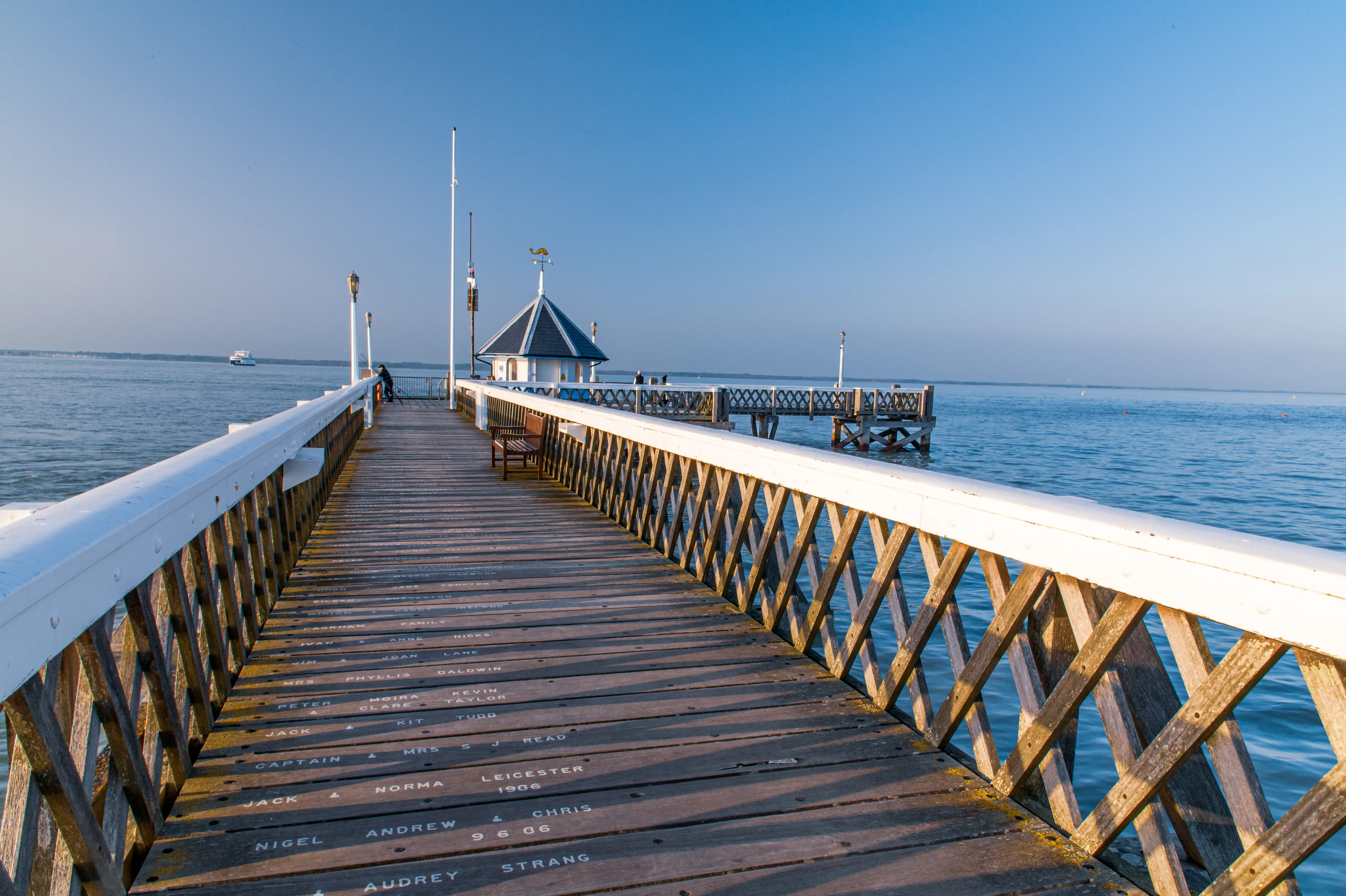 The image depicts a long wooden pier extending into a body of water, likely the ocean or a large lake. The pier has a white railing and is adorned with names inscribed on the wooden planks. There is a small gazebo-like structure with a pointed roof situated on the pier. The sky is clear and blue, indicating a sunny day, and the water appears calm. A few people can be seen on the pier, enjoying the serene environment.
