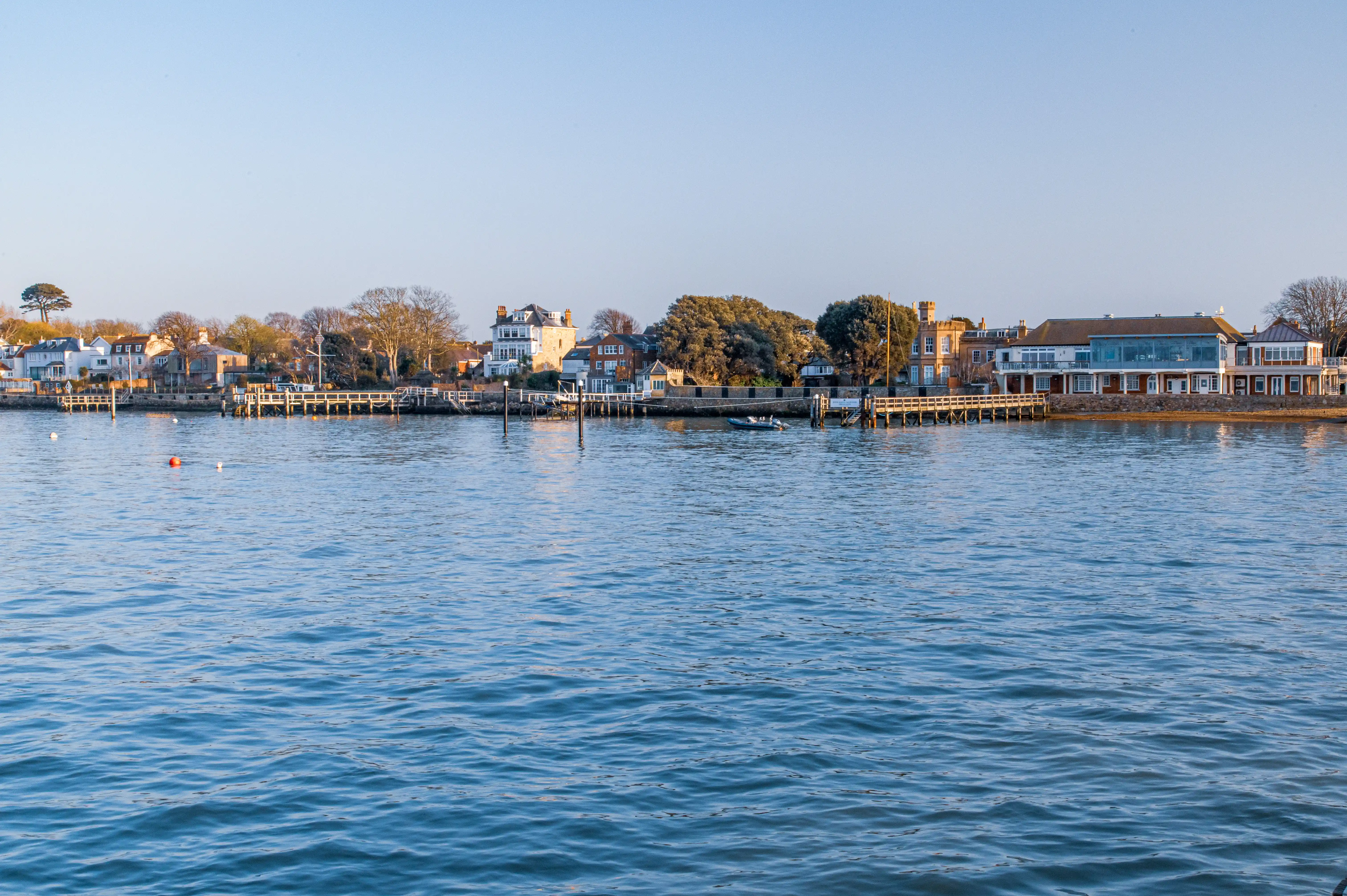 The image depicts a serene waterfront scene with a variety of residential buildings and houses lining the shore. There is a wooden pier extending into the water, and a small boat is visible near the pier. The water is calm, reflecting the clear sky above. The overall atmosphere is peaceful, suggesting a quiet, possibly coastal town.