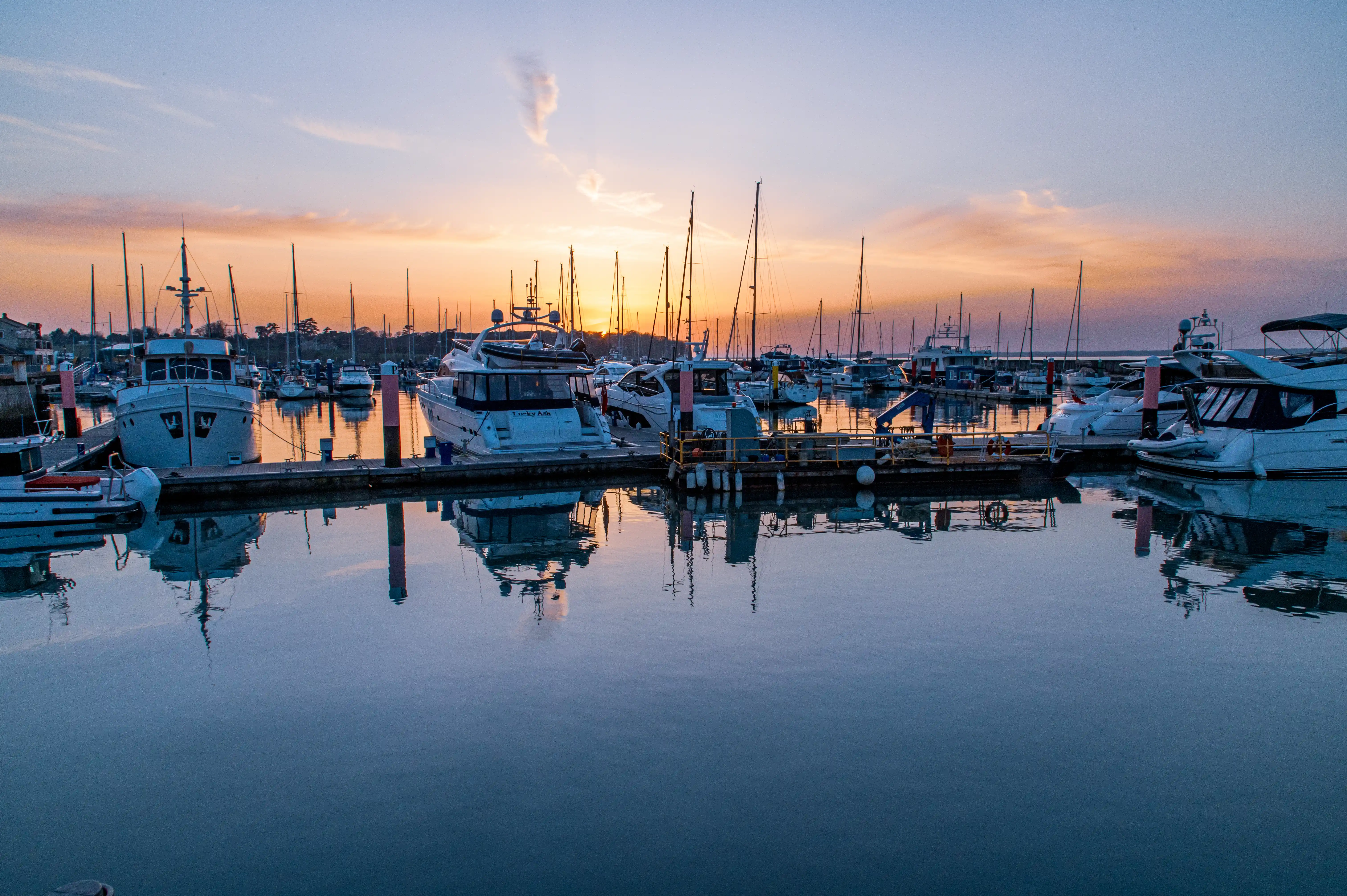 The image depicts a serene marina at sunset. Several boats and yachts are docked in calm water, creating a peaceful reflection. The sky is painted with hues of orange and pink, indicating the sun is setting. The masts of the boats are silhouetted against the colorful sky, adding to the tranquil ambiance of the scene.