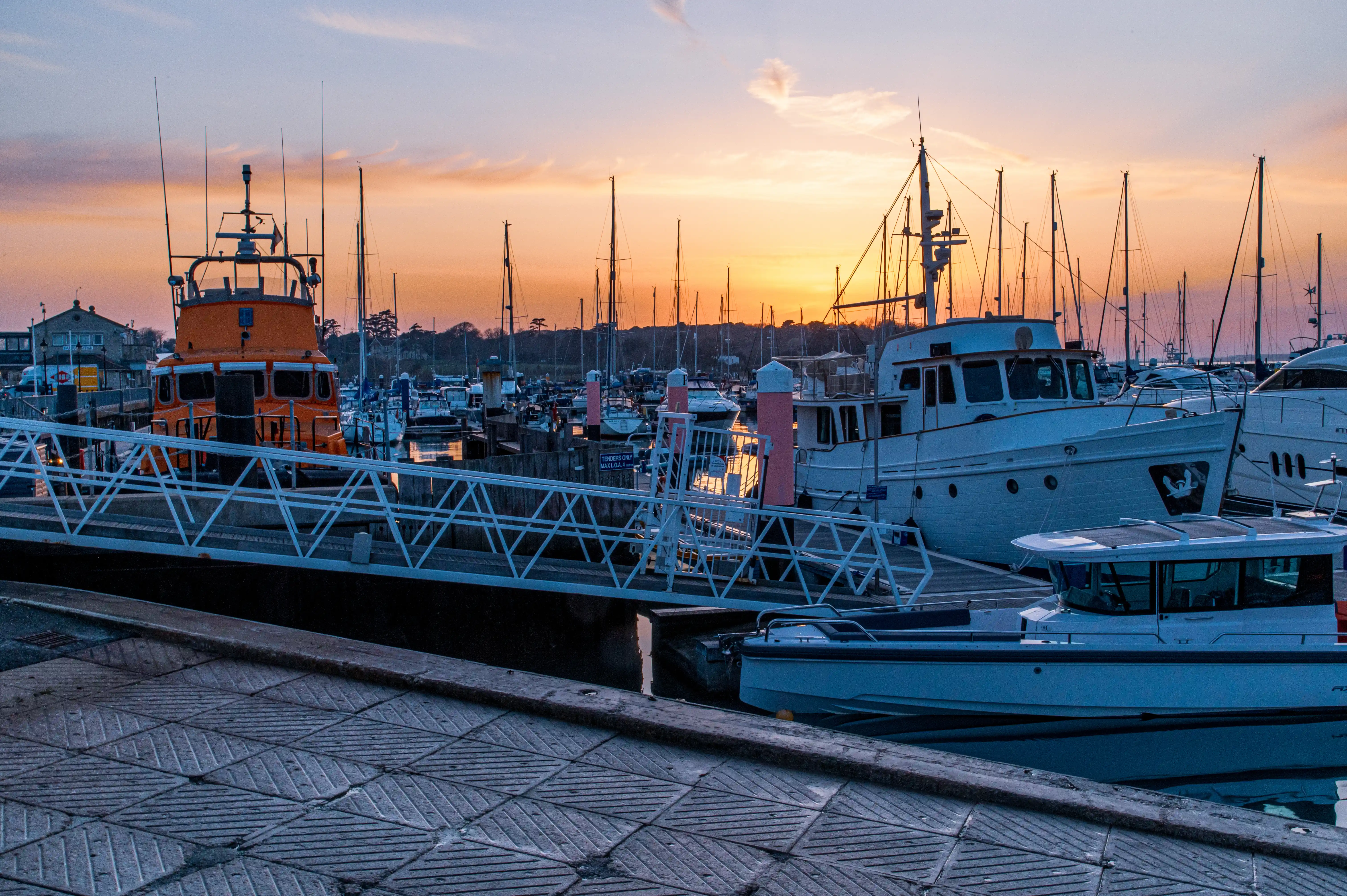 The image depicts a serene marina at sunset. Various boats and yachts are docked, with their masts silhouetted against the colorful sky. A prominent orange boat is visible on the left side of the image. The scene is calm and picturesque, with the setting sun casting a warm glow over the entire harbor.
