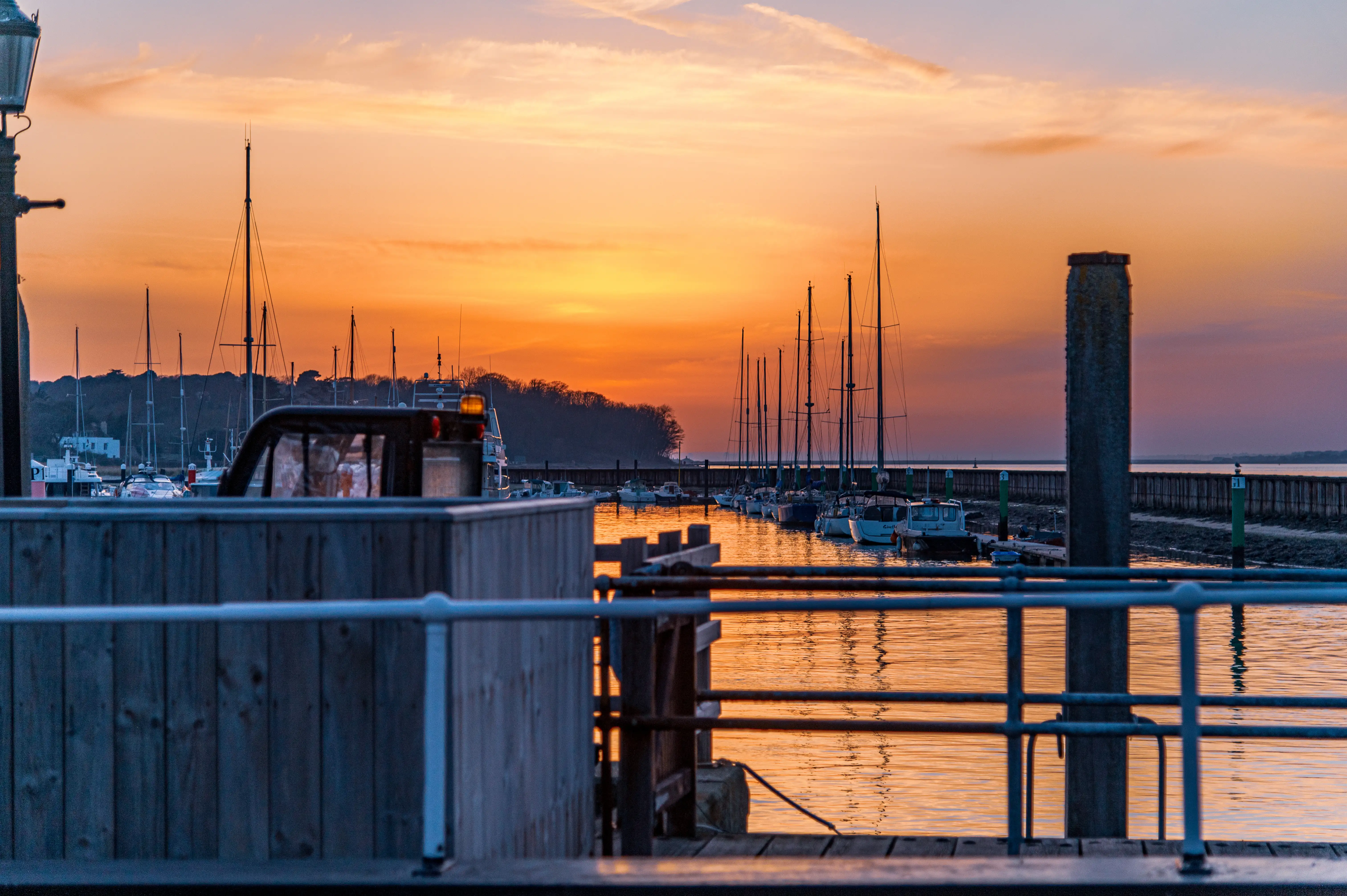 The image depicts a serene marina at sunset. The sky is painted with hues of orange and yellow, reflecting on the calm water. Several sailboats and yachts are docked in the harbor, their masts silhouetted against the vibrant sky. The foreground features a wooden railing and a partially visible vehicle, adding a touch of everyday life to the tranquil scene.