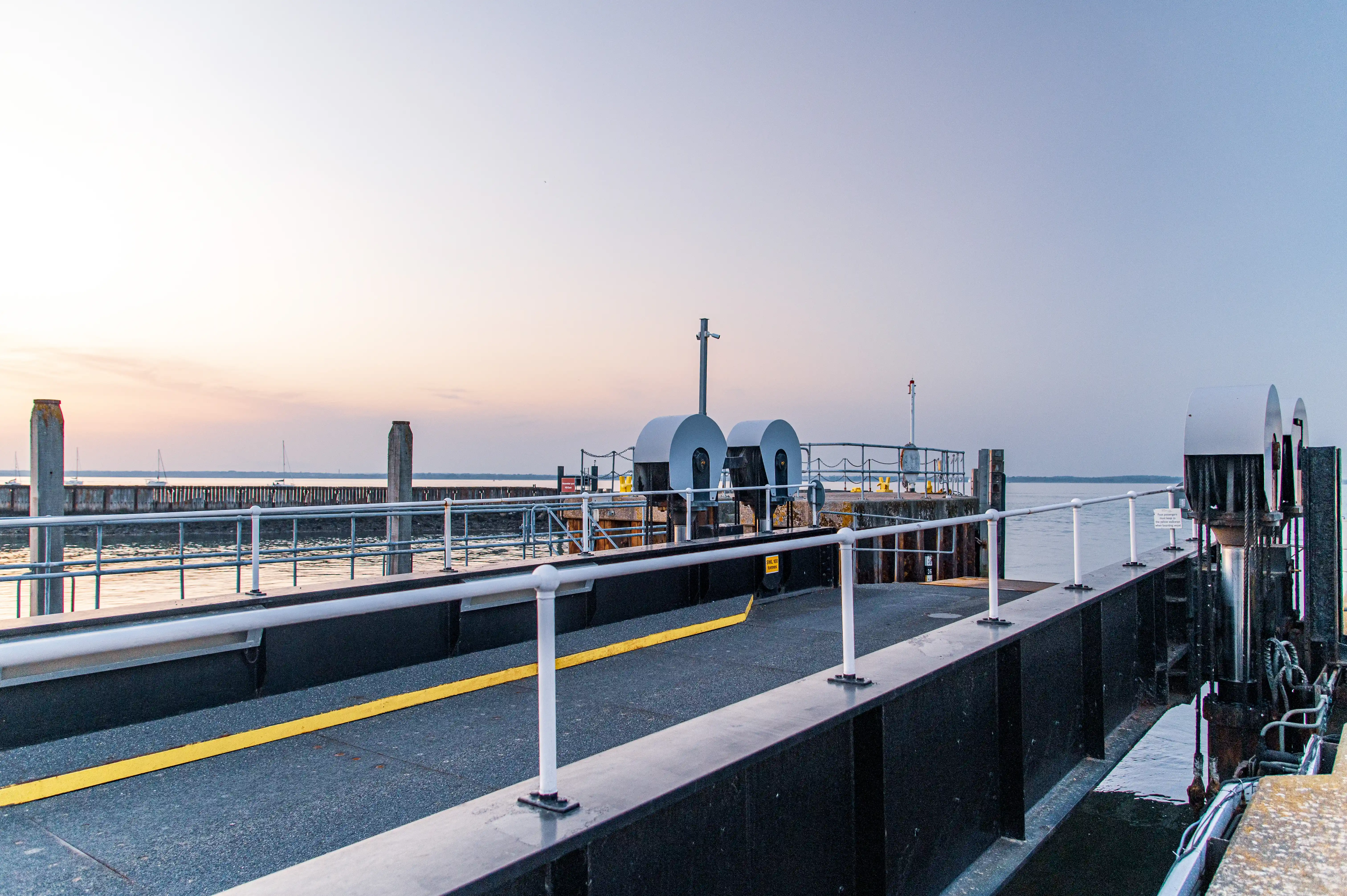 The image depicts a dock or pier extending into a body of water during what appears to be either dawn or dusk. The dock is equipped with railings and various mechanical structures, likely used for mooring boats. The sky is clear with a soft gradient from light to darker hues, suggesting calm weather conditions. The water is relatively calm, with a few distant sailboats visible on the horizon.