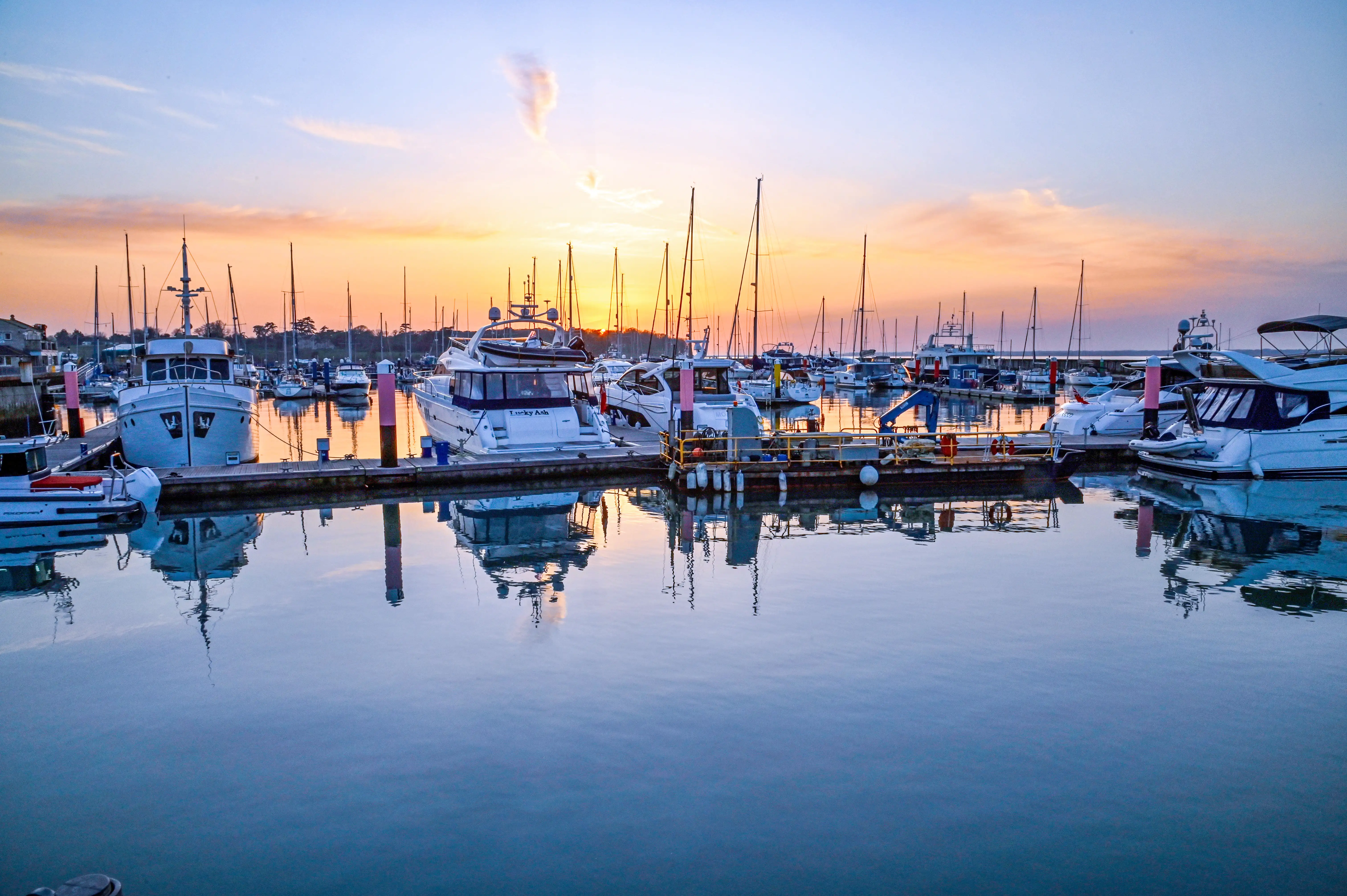 The image depicts a serene marina at sunset. Several boats and yachts are docked in calm waters, creating a picturesque reflection. The sky is painted with hues of orange and pink, indicating the sun is setting. The scene is tranquil and visually appealing, showcasing the beauty of the marina during the evening.