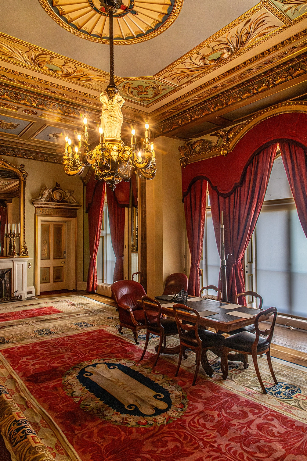The image depicts an ornately decorated room with intricate ceiling designs and a grand chandelier. The room features red curtains, antique furniture including chairs and a table, and a detailed, colorful rug. The walls are adorned with decorative elements, and there is a fireplace visible in the background. The overall ambiance suggests a historical or luxurious setting.