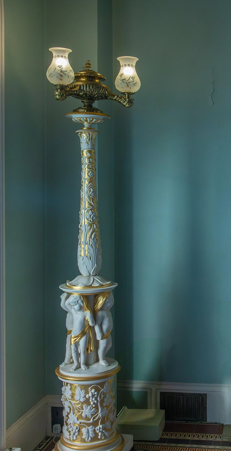 The image depicts an ornate, antique floor lamp. The lamp features a tall, intricately decorated column with gold accents and white detailing. At the base, there are sculpted figures of cherubs draped in gold cloth. The lampshades are frosted with a floral design, and the lamp is set against a teal-colored wall.