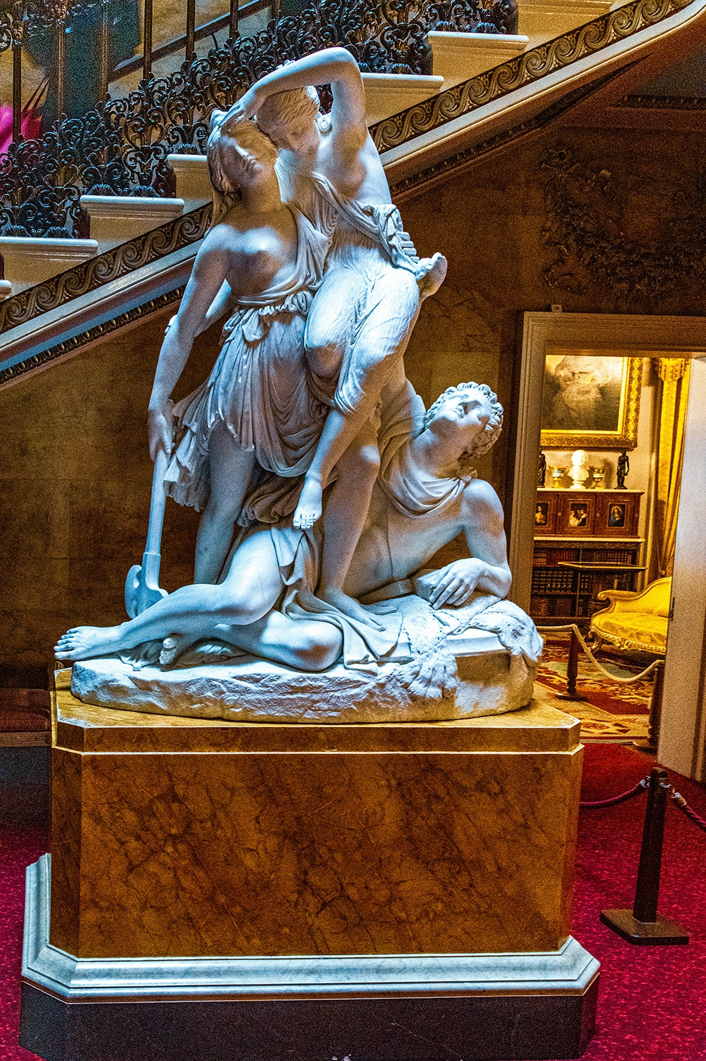 The image depicts a classical marble sculpture situated in an ornate indoor setting. The sculpture features three human figures: a female figure standing and holding an object, another female figure draped over her shoulder, and a male figure kneeling and appearing to be in distress. The setting includes a richly decorated staircase, a warmly lit room with bookshelves, and a red carpeted floor