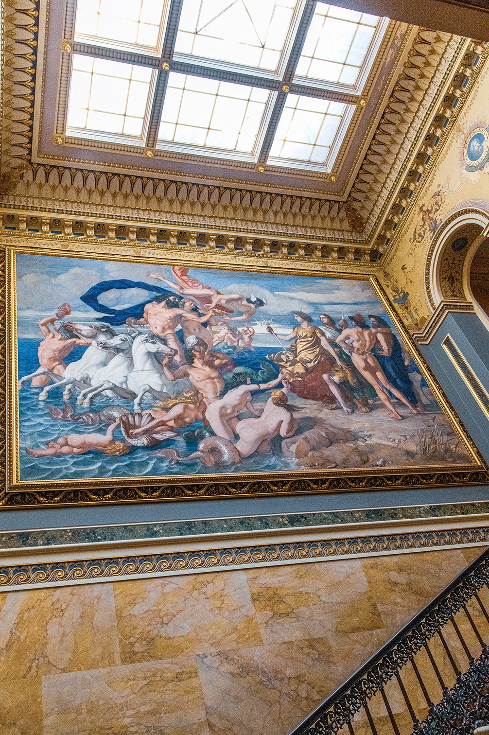 The image depicts an ornate interior space featuring an elaborate ceiling mural. The mural showcases a classical scene with mythological figures, including humans and sea creatures, set against a seascape background. The architectural details include intricate moldings, a skylight, and marble walls, suggesting a grand and historic building, possibly a museum or a palace.