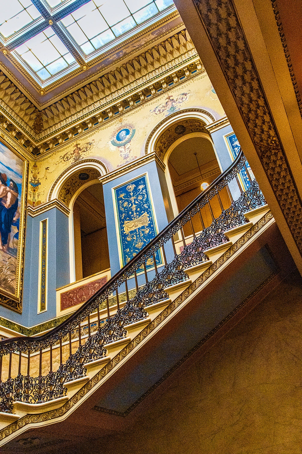 The image depicts an ornate interior of a grand building, featuring a beautifully decorated staircase with intricate wrought iron railings. The walls and ceiling are adorned with elaborate frescoes and detailed moldings, showcasing classical art and architectural elements. The staircase curves gracefully, leading to an upper level with arched openings and additional artwork. The overall ambiance is one of opulence and historical elegance, likely indicative of a palace or a significant public building.