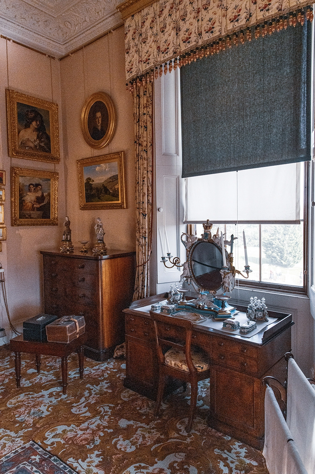 The image depicts a vintage, elegantly decorated room. It features antique wooden furniture, including a dresser with a mirror and a small table. The walls are adorned with several framed paintings and a decorative cornice. The room has a patterned carpet and a window with dark roller blinds, allowing some natural light to enter.