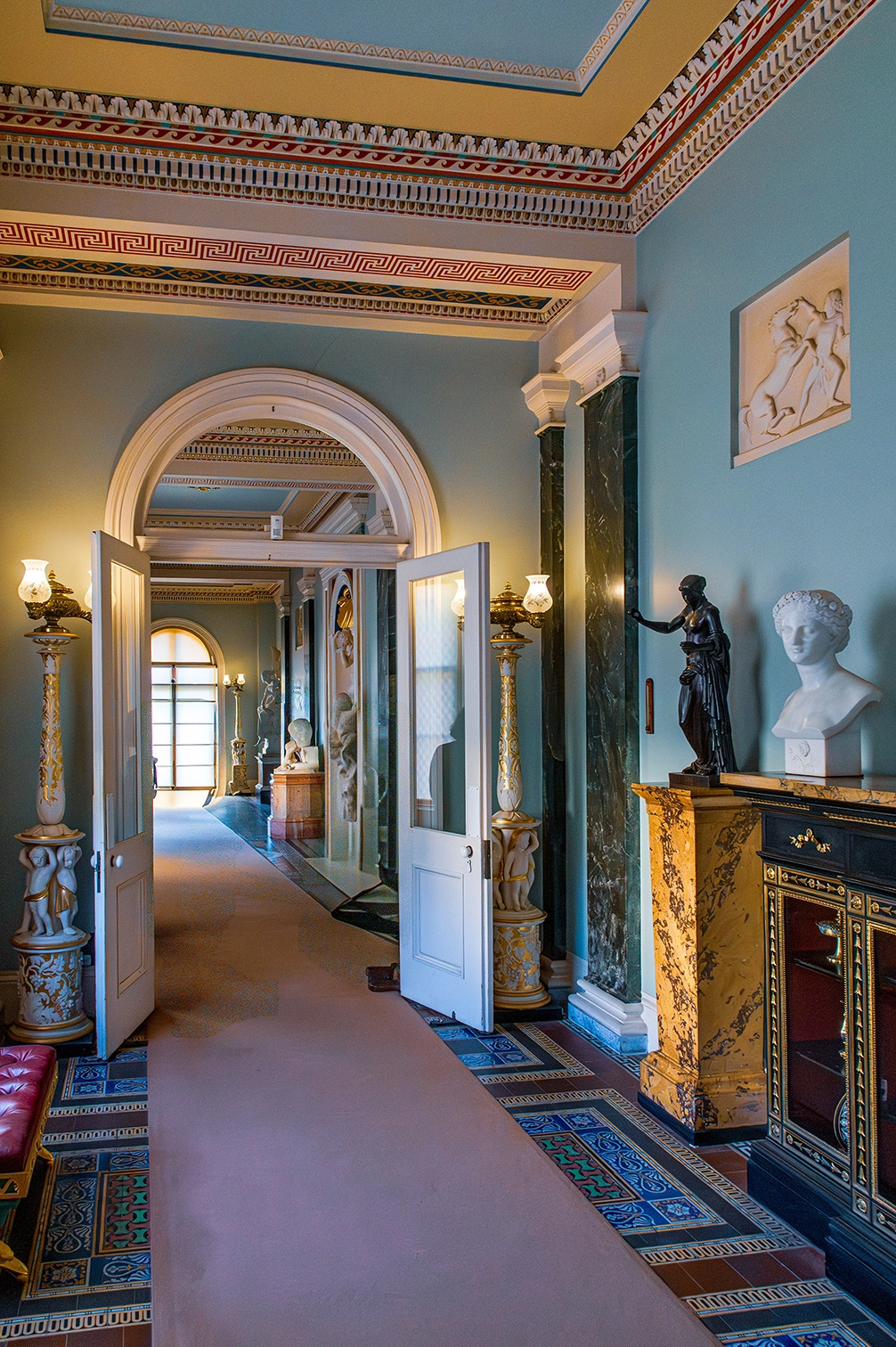 The image depicts an ornate hallway in what appears to be a historic or grand building. The hallway features classical architectural elements such as arches, columns, and intricate ceiling moldings. It is adorned with various statues and sculptures, including a bust on a marble-topped cabinet and a dark statue of a figure. The walls are painted in a light blue-green color, and the floor is covered with a patterned carpet and decorative tiles. The space is illuminated by elegant wall-mounted lamps.