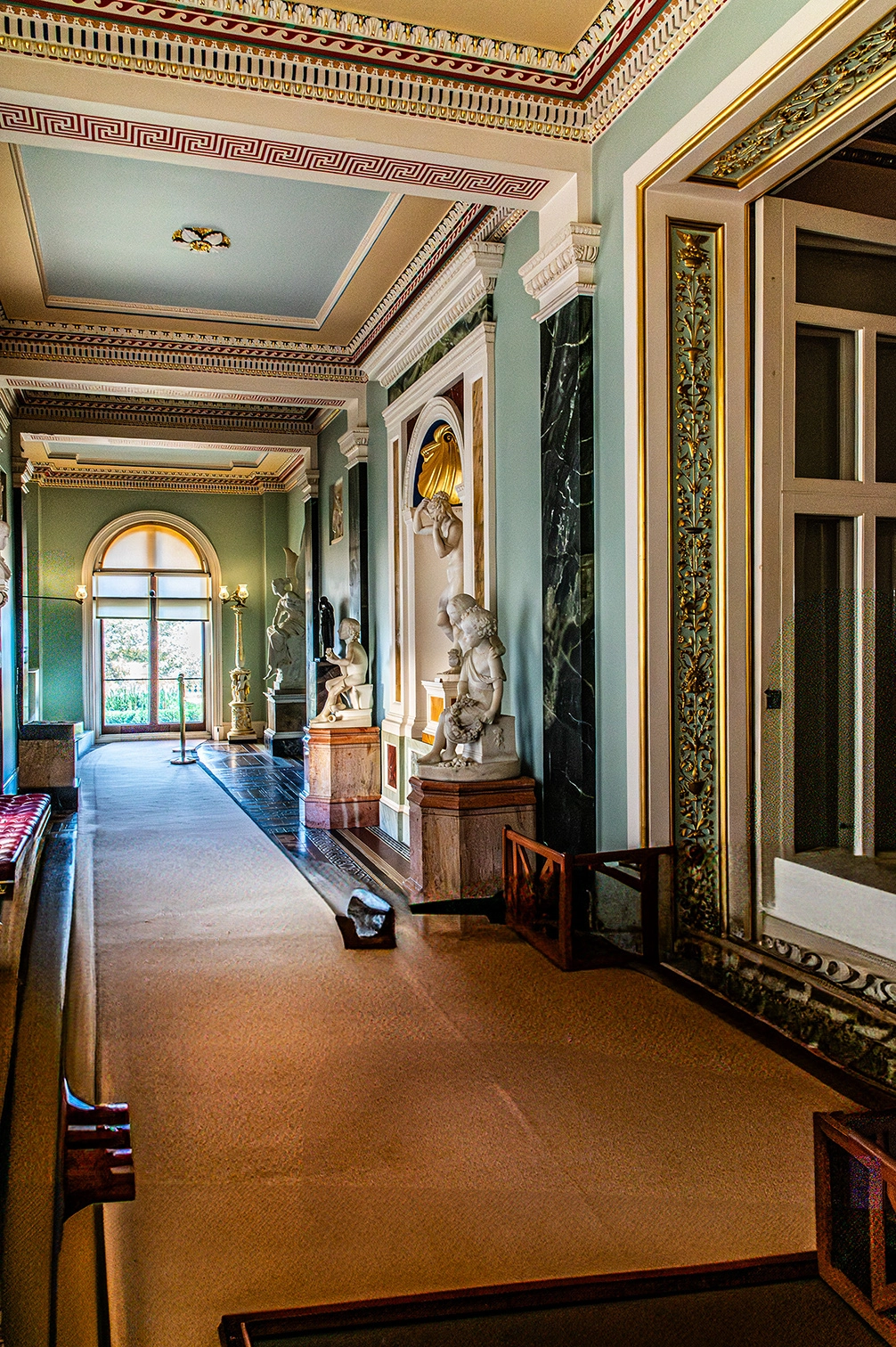 The image depicts an ornate hallway with classical architectural elements. It features intricately designed ceilings, marble columns, and several statues placed on pedestals along the corridor. The walls are adorned with decorative moldings and gold accents. The hallway is carpeted and leads to a large arched window at the far end, allowing natural light to illuminate the space.