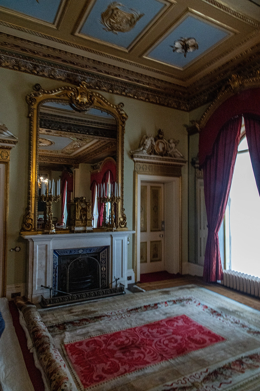 The image depicts an elegantly decorated room featuring a large, ornate mirror above an intricately designed fireplace. The room has a classic and luxurious aesthetic with detailed moldings on the ceiling and walls. Heavy red curtains frame a window, and a large, patterned rug covers the floor. The overall ambiance suggests a historical or period-style interior design.