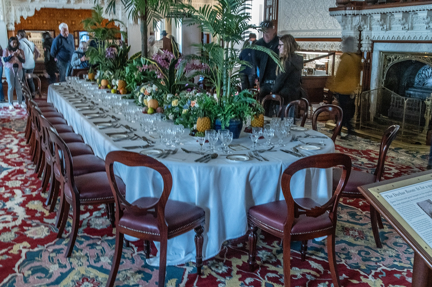 The image depicts a large, elegantly set dining room with a long table adorned with white tablecloths, numerous wine glasses, plates, and cutlery. The table is decorated with tropical plants and flowers, including pineapples. The room features ornate decor, including a patterned carpet and a grand fireplace. Several people are present, some observing the setup and others casually interacting in the background.