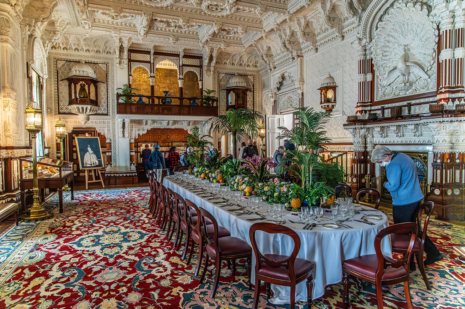 The image depicts an ornately decorated dining room with intricate architectural details and elaborate furnishings. A long table is set with white tablecloths, glassware, and floral centerpieces, surrounded by wooden chairs. Several people are present, some attending to the table settings. The room features high, detailed ceilings, large paintings, and a richly patterned carpet. The ambiance suggests a formal and luxurious setting, possibly in a historic or culturally significant building.