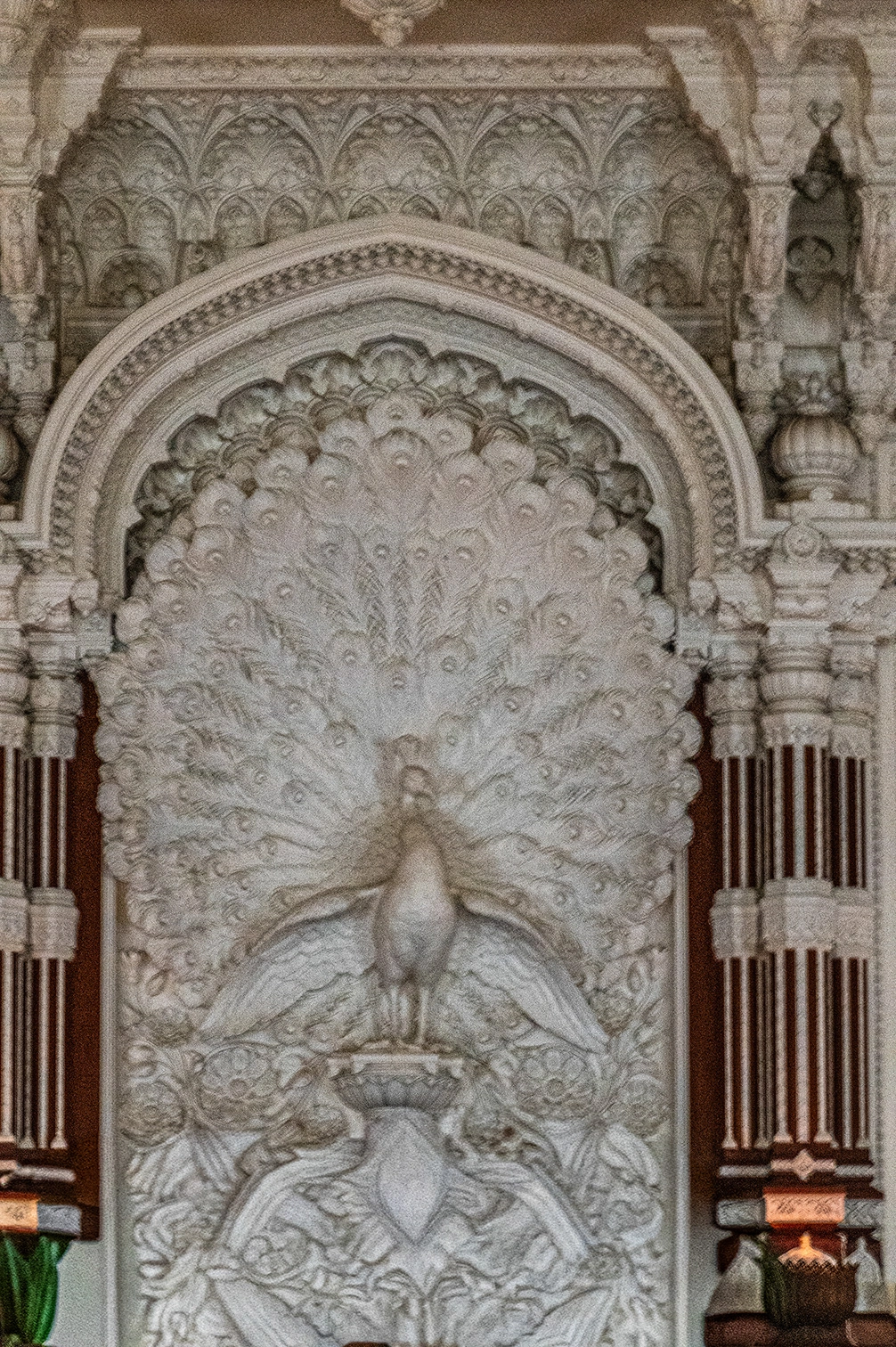The image depicts an intricately carved stone archway featuring a peacock with its feathers fully displayed. The arch is adorned with elaborate patterns and surrounded by detailed architectural elements, including columns and decorative motifs. The craftsmanship highlights fine details, particularly in the feathers and surrounding foliage. The overall design suggests a blend of artistic and architectural styles, possibly indicative of a historical or cultural monument.