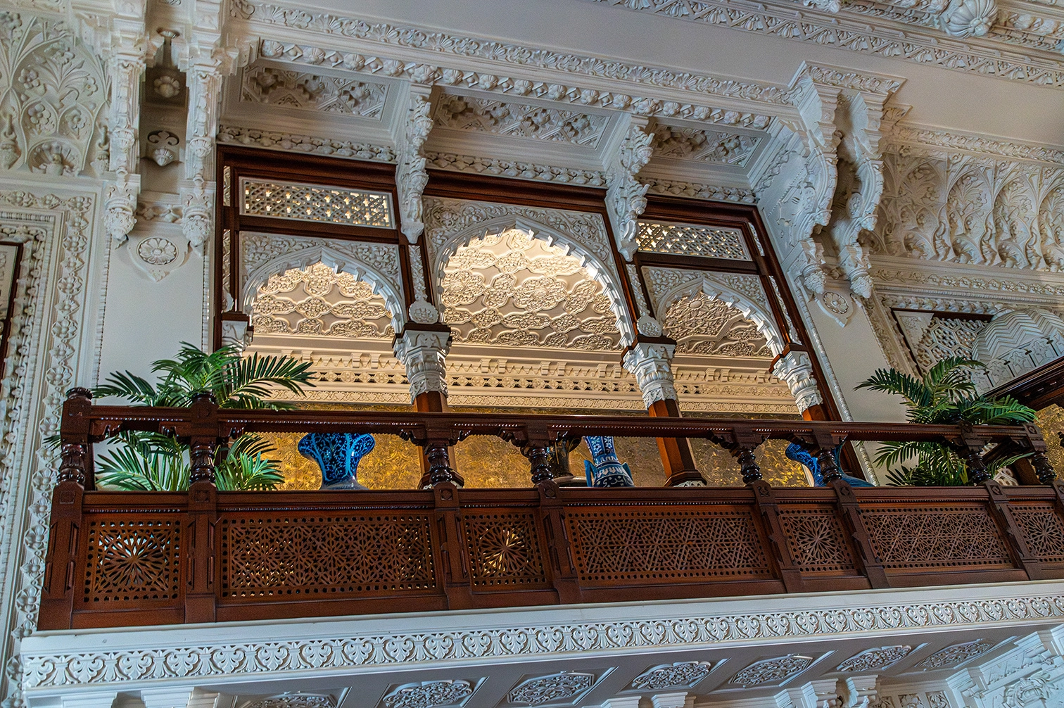 The image depicts an intricately designed interior space with elaborate architectural details. The upper section features ornate carvings and decorative arches, while the lower section includes a wooden balcony adorned with detailed latticework. The balcony holds potted plants and decorative vases, contributing to the overall aesthetic. The craftsmanship showcases a blend of intricate patterns and fine detailing, reflecting a sophisticated and elegant architectural style.